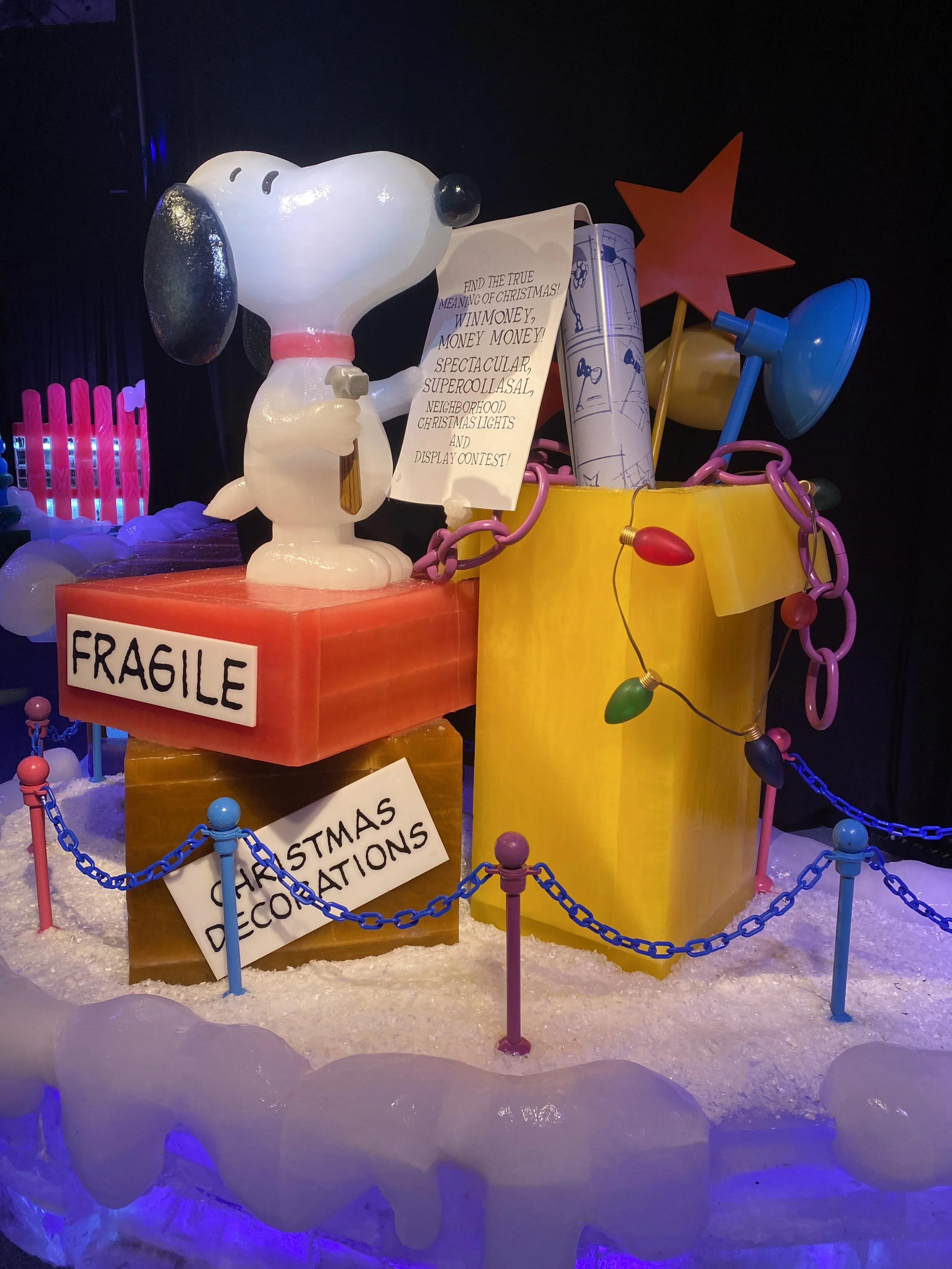 Snoopy figure holding a paper with Christmas-themed text, surrounded by colorful holiday decorations and a sign that reads 'Fragile'.