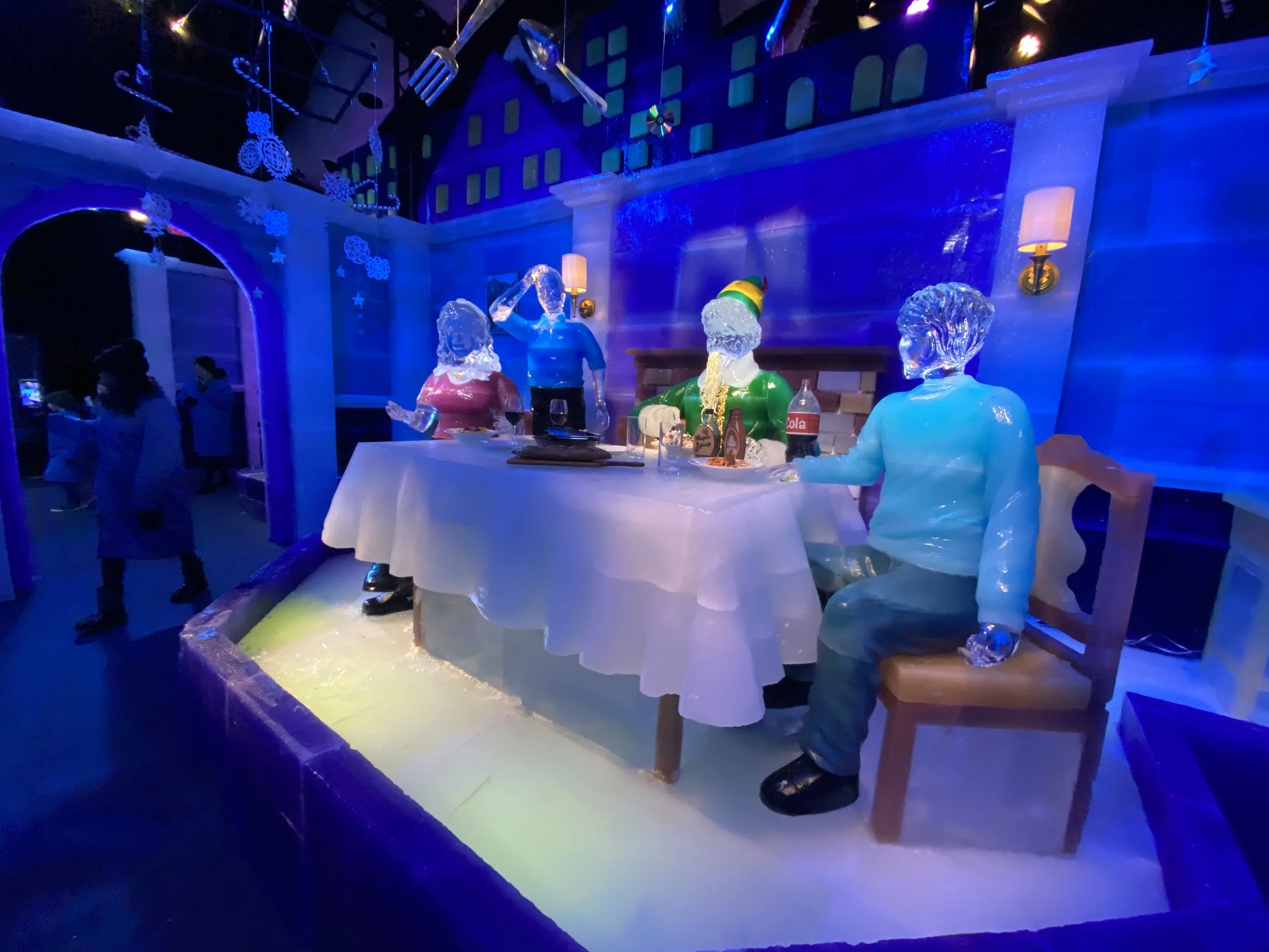 Ice sculptures of four people at a restaurant or ice bar, with a snowman-like figure dressed in a green and yellow hat and a purple shirt. The sculptures are illuminated in colorful blue and purple lights, with kitchen utensils and snowflake decorati