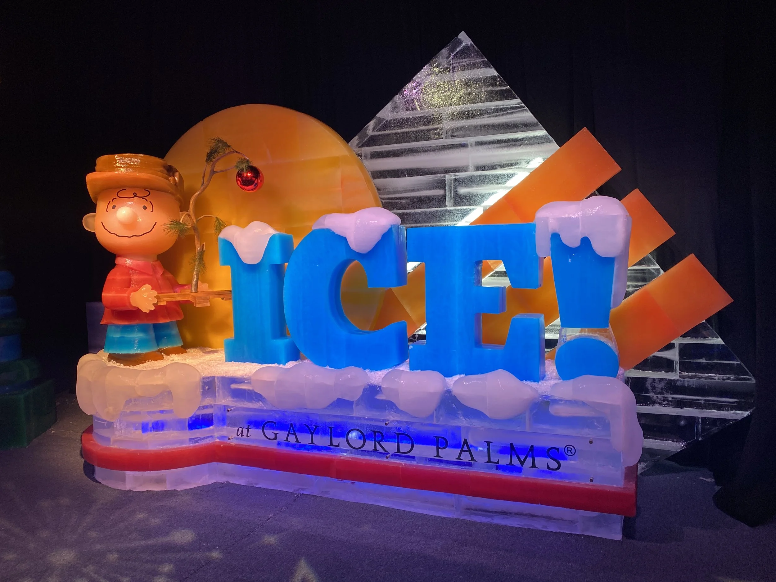 Ice sculpture of Charlie Brown holding a small Christmas tree with a red ornament. The sculpture is illuminated with colorful lights and has the text 'at GAYLORD PALMS' at the base.