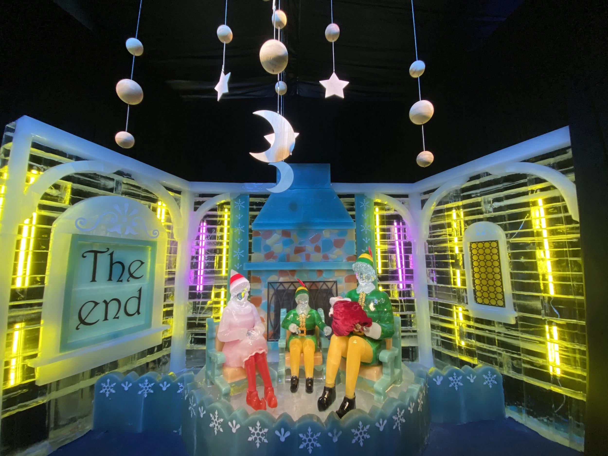 Colorful Christmas display with illuminated hanging moons, stars, and planets, and holiday-themed figurines sitting on a bench under a black ceiling.