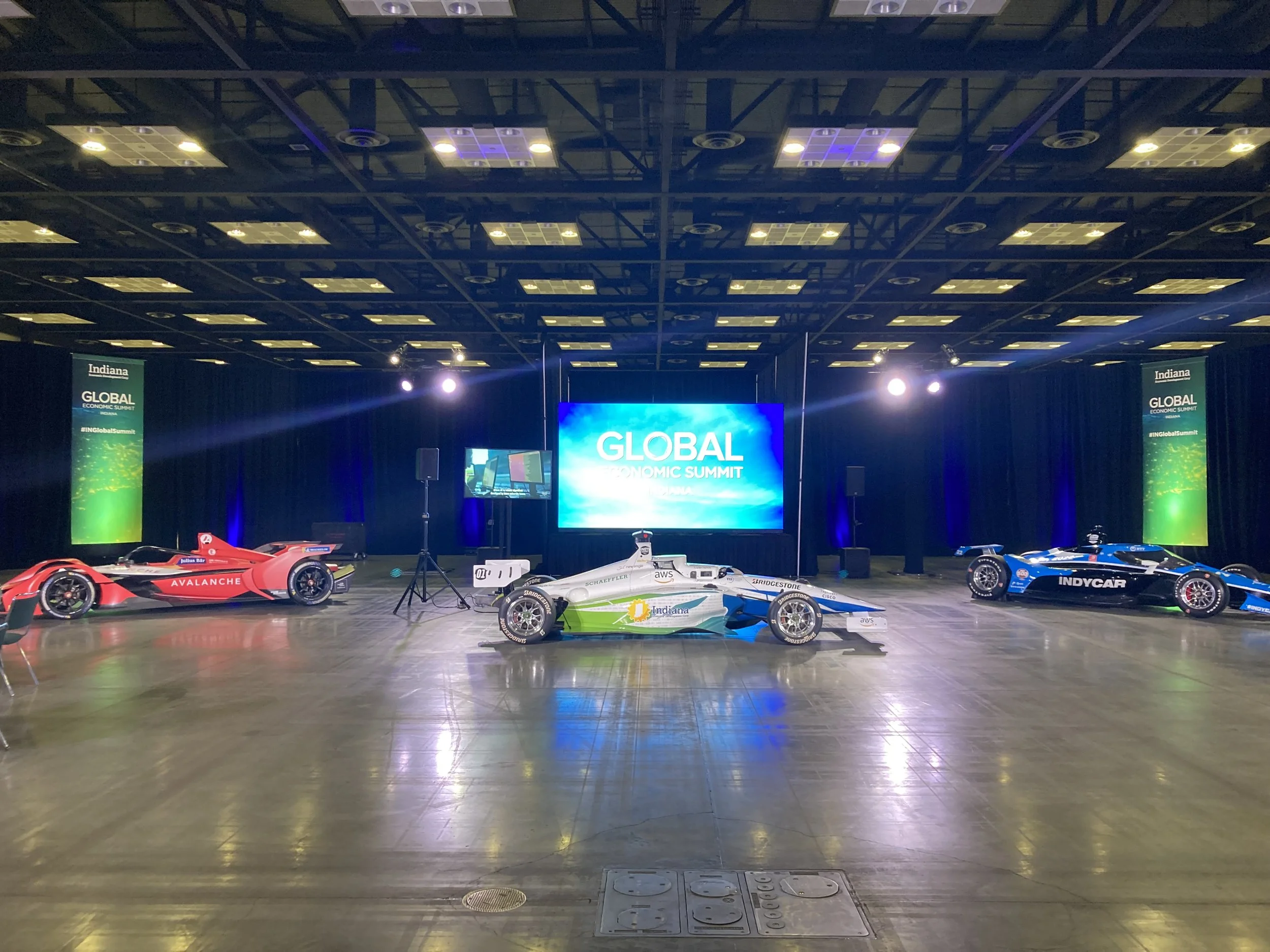 Indoor stage with race cars on display, large screen shows 'Global Economic Summit,' banners on each side read 'Indiana Global Economic Summit.' Overhead lights and a black ceiling.