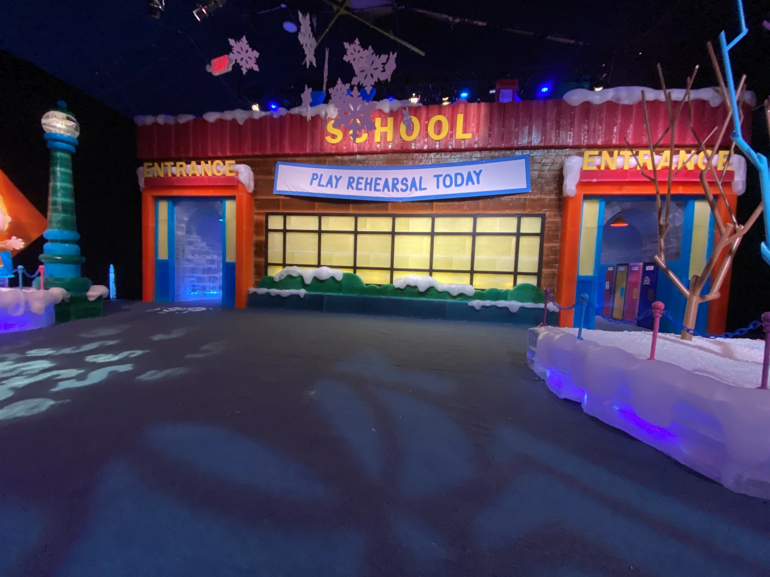 Indoor scene with a sign that reads 'SCHOOL,' decorated with snowflakes and snow-like material, indicating a winter theme or holiday event. There are colorful decorations, and a banner that says 'PLAY REHEARSAL TODAY.'