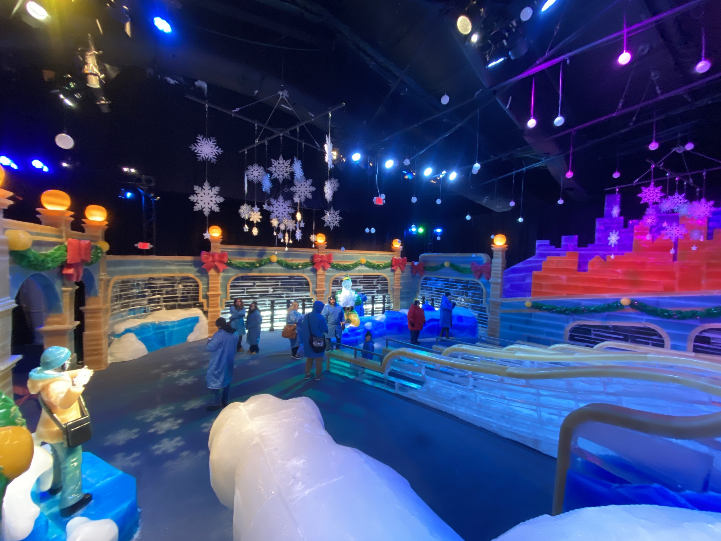 Interior of an ice skating rink decorated with Christmas-themed ornaments such as snowflakes, garlands, and bows. People are warming up or preparing to skate around the rink, with some sitting and others standing. The ceiling has colorful lighting ef