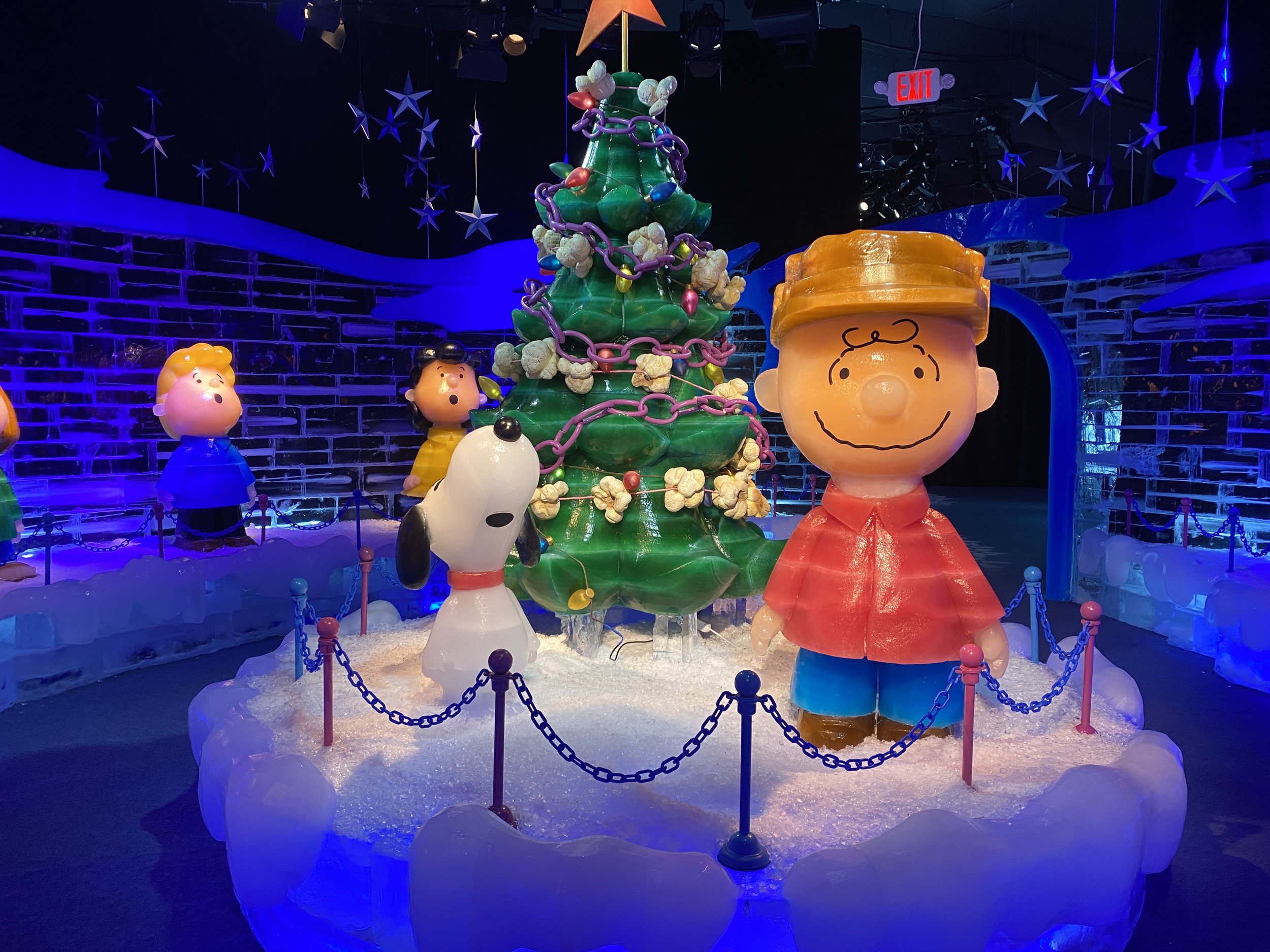 Colorful Peanuts Christmas tree with purple, pink, and white chains, surrounded by Peanuts characters Lucy, Charlie Brown, and Snoopy, in a festive indoor setting with blue lighting and star decorations.