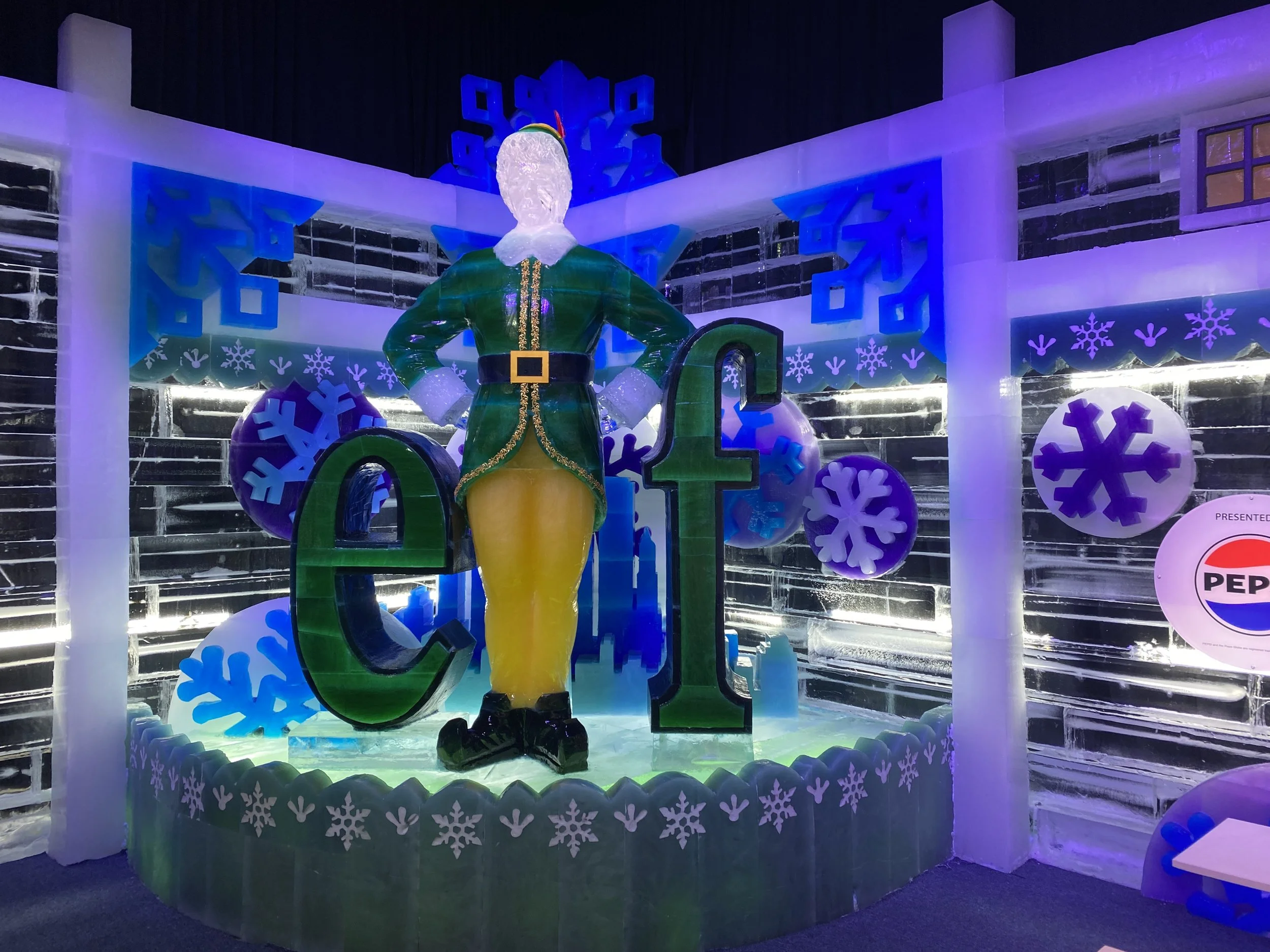 Ice sculpture of a person dressed as Santa Claus holding a cane, with large colorful numbers 20 behind, surrounded by ice snowflakes and decorations, illuminated by blue and purple lights.