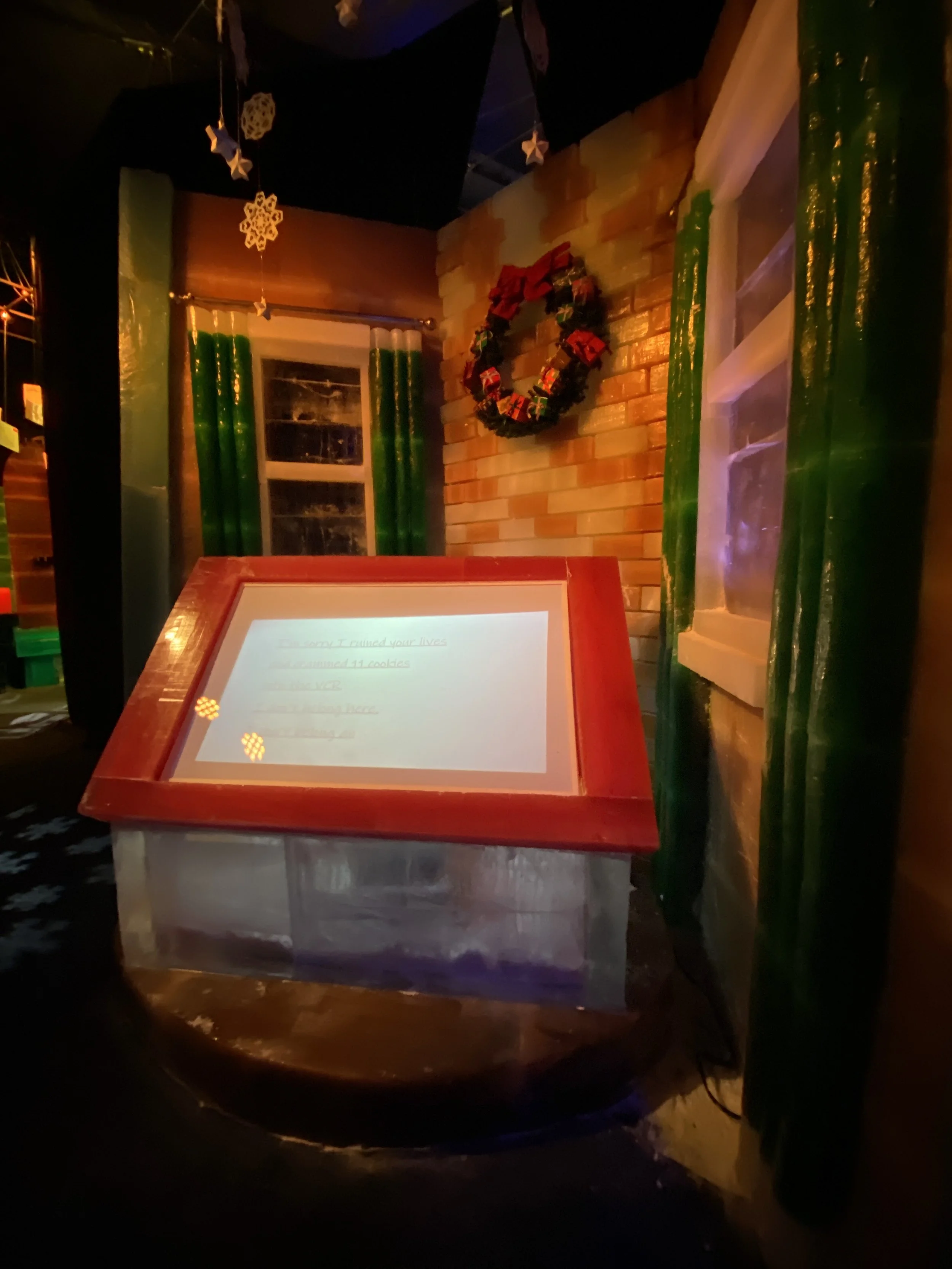 A Christmas wreath decorated with small presents hangs on a brick wall. There are green curtains on either side of a window with white trim. A blog or menu is displayed on a red-framed stand in the foreground. Star-shaped and snowflake ornaments hang