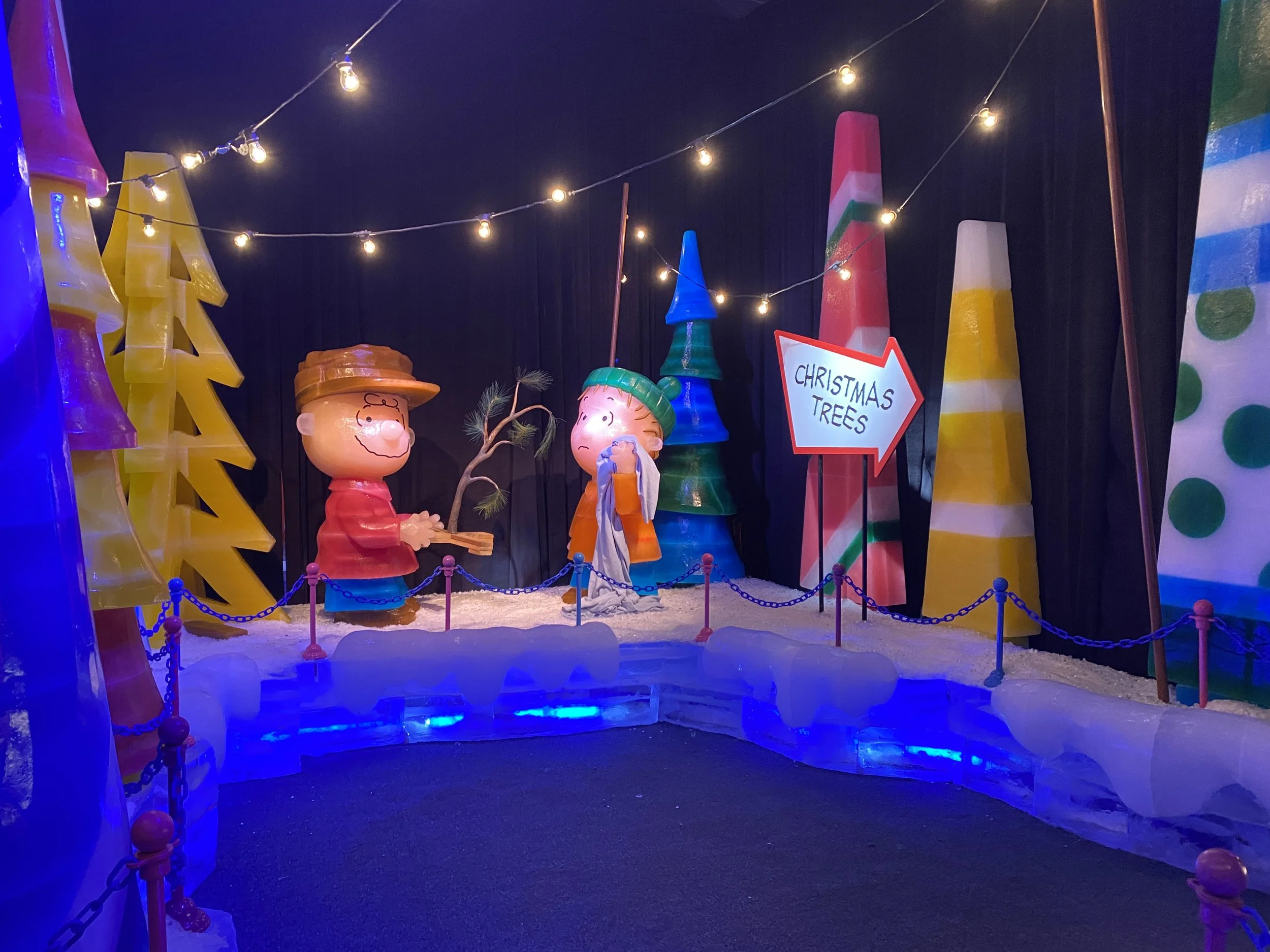 Decorative display of cartoon-like characters and oversized Christmas trees with string lights and a sign pointing to Christmas trees.