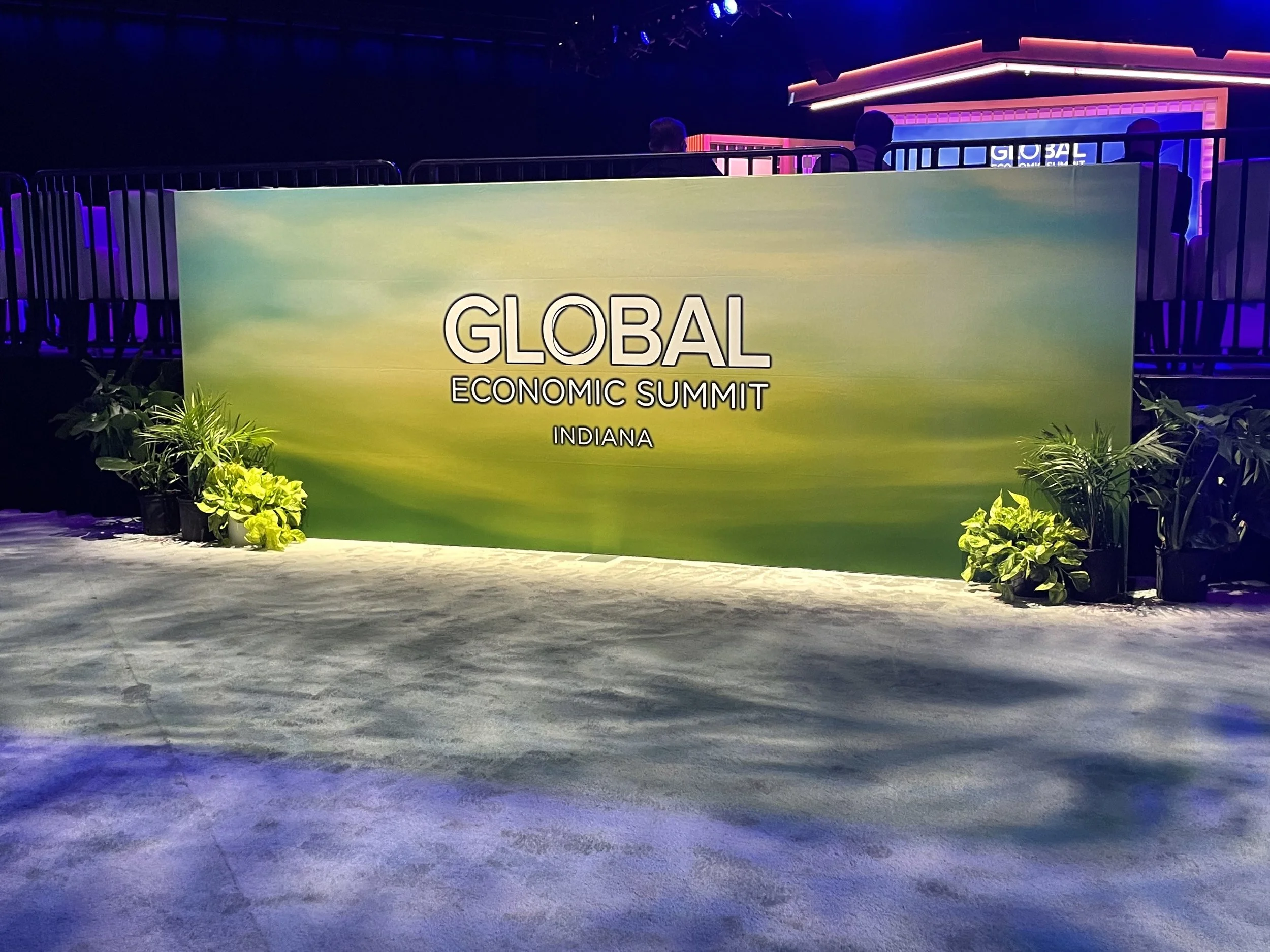 A large green sign with white text reading 'GLOBAL ECONOMIC SUMMIT INDIANA' in front of a stage with purple and pink lighting, surrounded by potted plants.