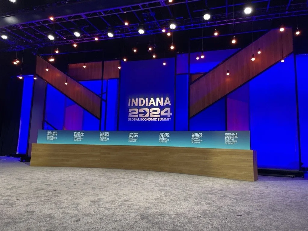 Stage set for the Indiana 2024 Global Economic Summit with a large screen displaying the event name and a long wooden desk in front. The backdrop features blue and purple lighting with hanging lights overhead.