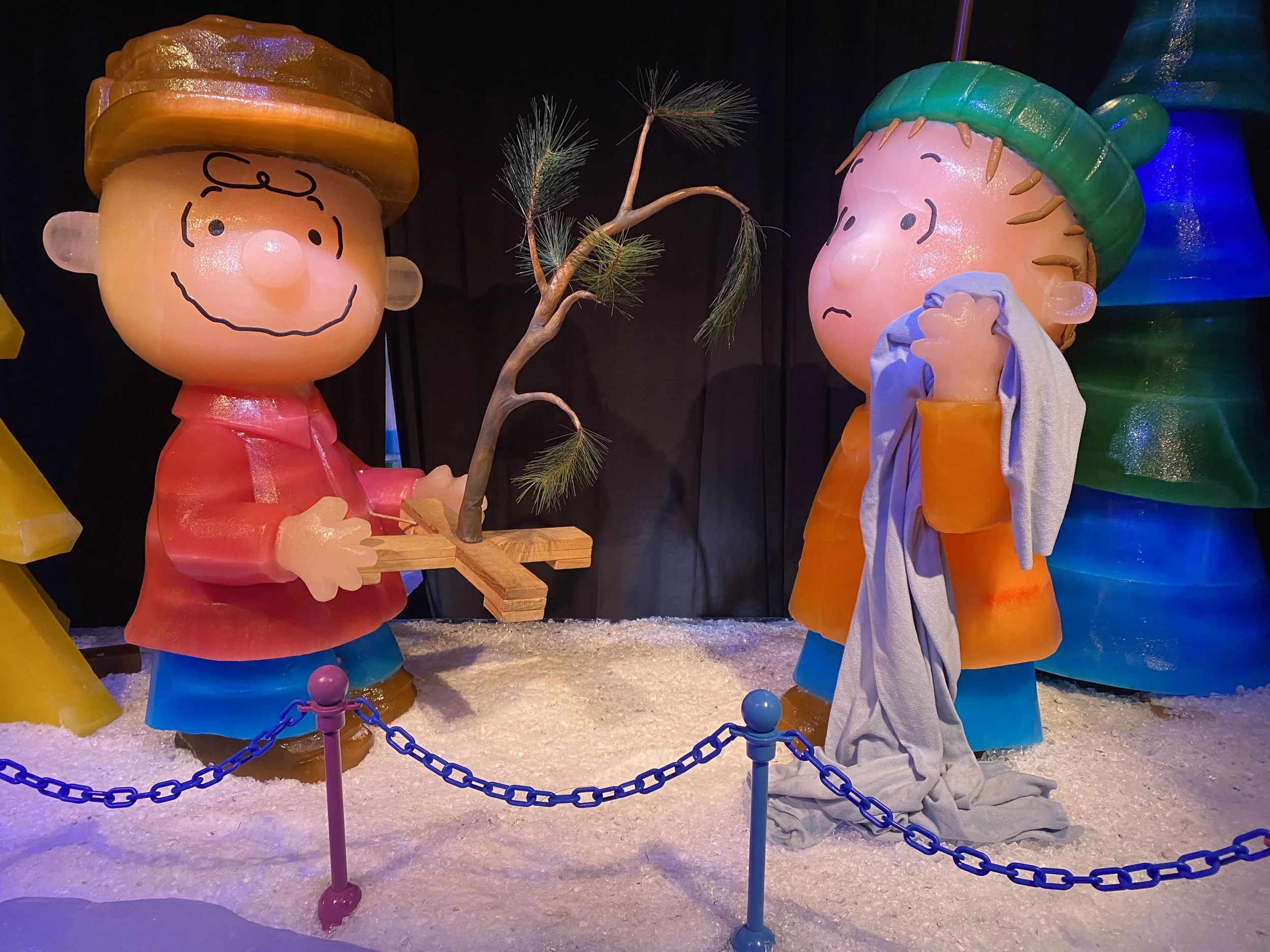 Winter scene with large, colorful comic-style characters of a boy with a yellow hat and a girl with a green hat, standing on snow with a small tree between them. The characters are part of a decorative display with a blue chain barrier in the foregro