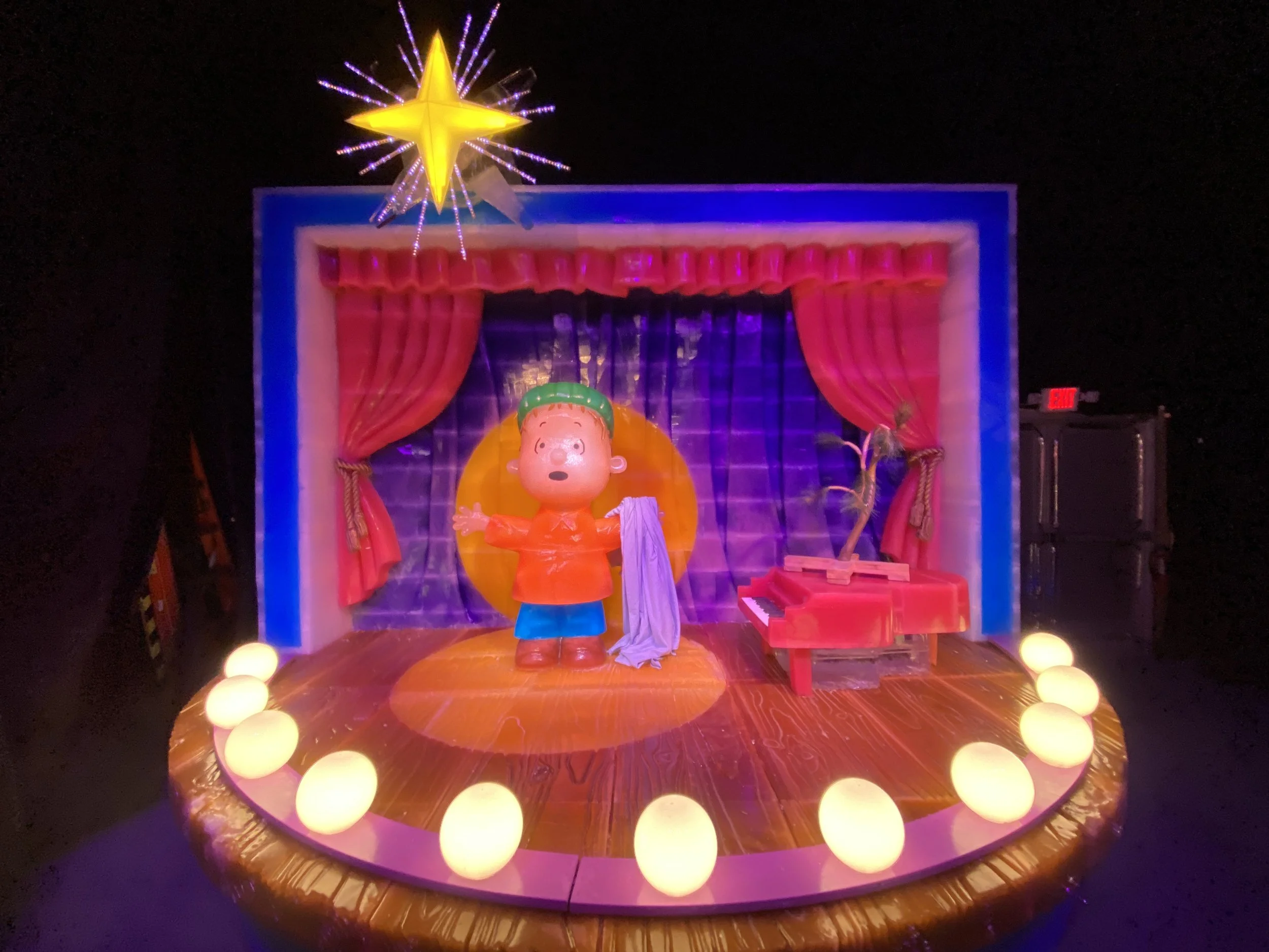 A colorful display with a toy character in front of a backdrop with red curtains and string lights, a large yellow star with purple and blue light accents at the top, and spherical lights encircling the base of the display.
