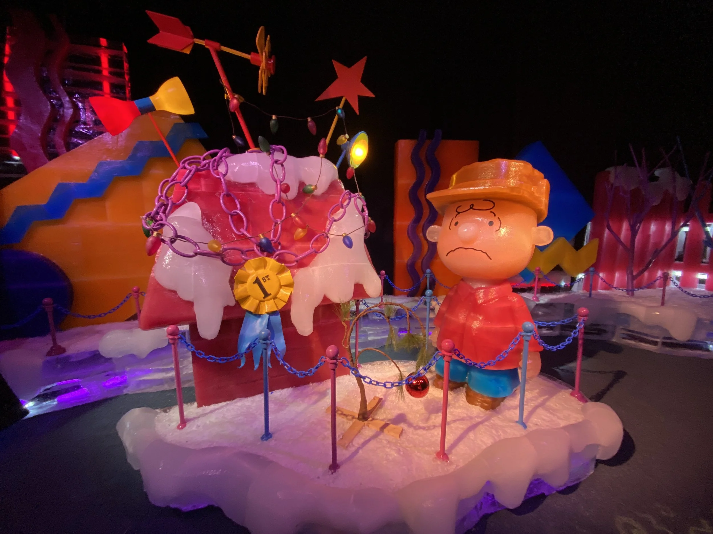 Colorful display featuring cartoon characters of Charlie Brown and Snoopy from Peanuts, decorated with string lights, stars, and holiday decorations, illuminated at night.