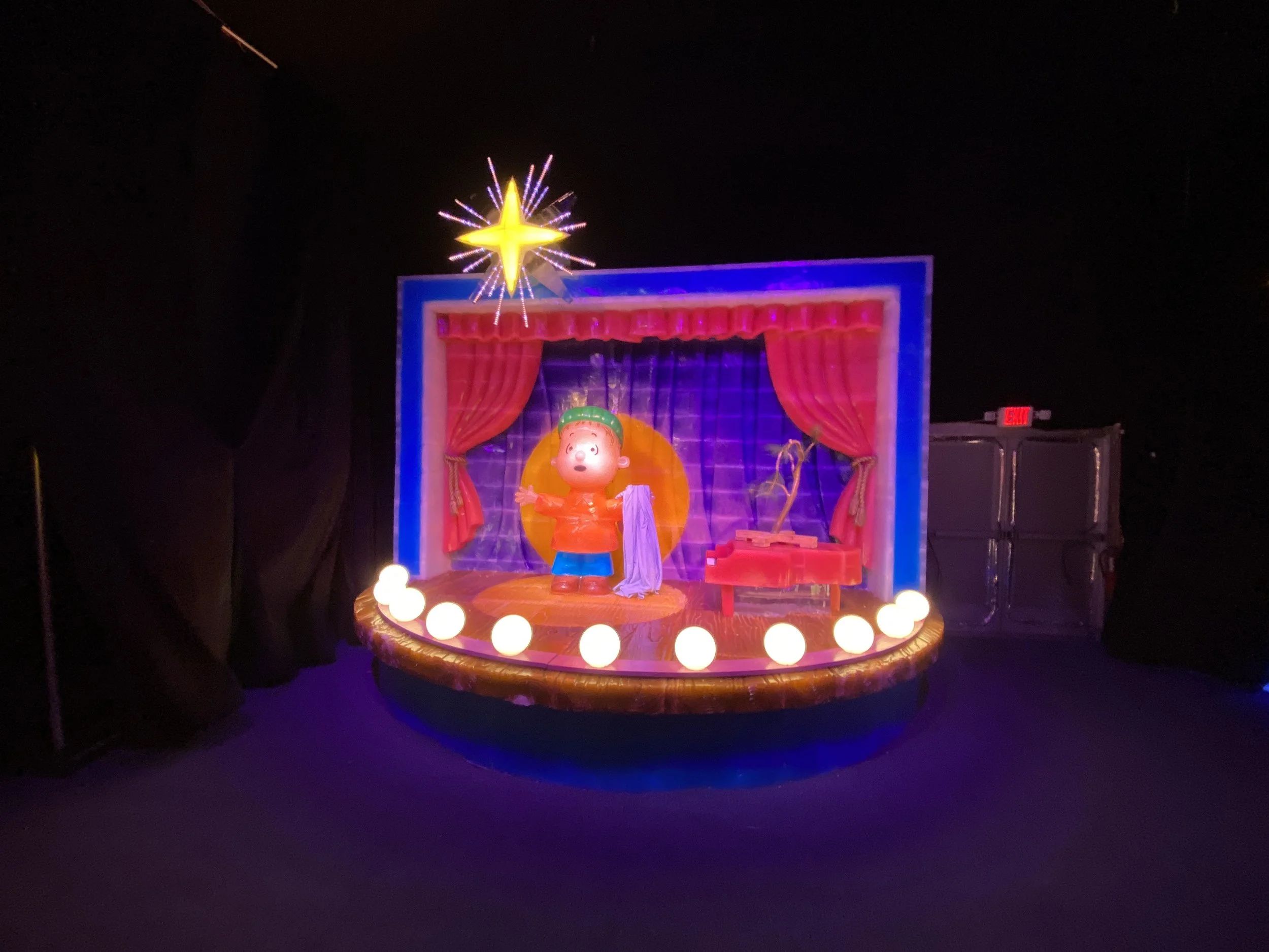 Colorful carnival or amusement park display with a cartoon character figure, red curtains, and decorative lights at night, topped by a star-shaped firework or light explosion.