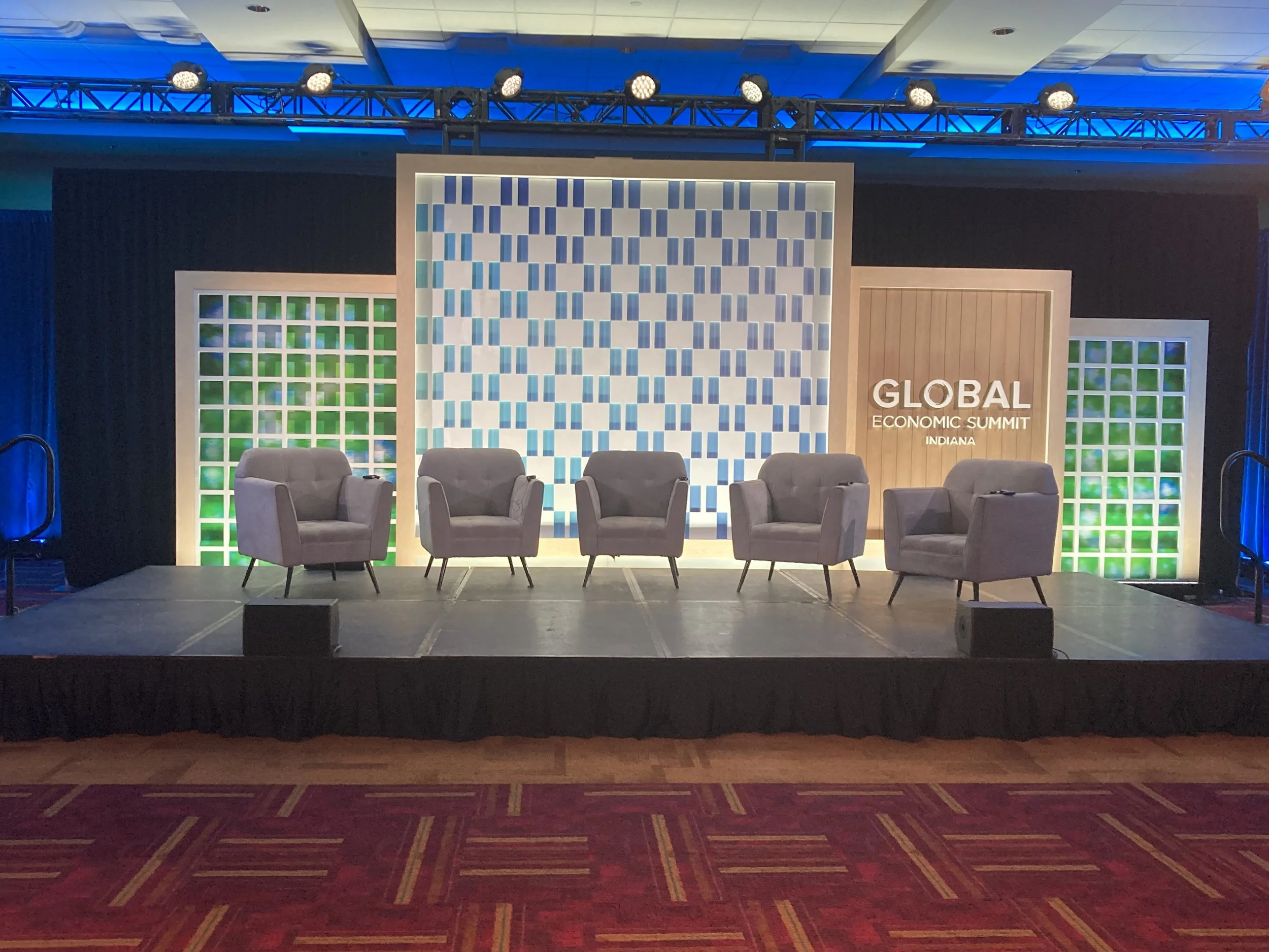 Stage setup at the Global Economic Summit in Indiana featuring three gray armchairs, a backdrop with blue and green geometric patterns, and a sign with the event name.