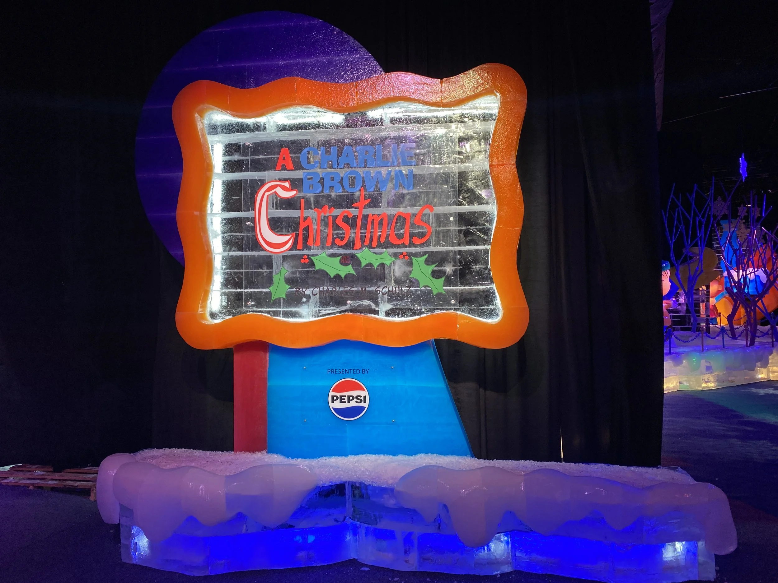 Ice sculpture with the text 'A Charlie Brown Christmas' and holly decorations, illuminated at night.