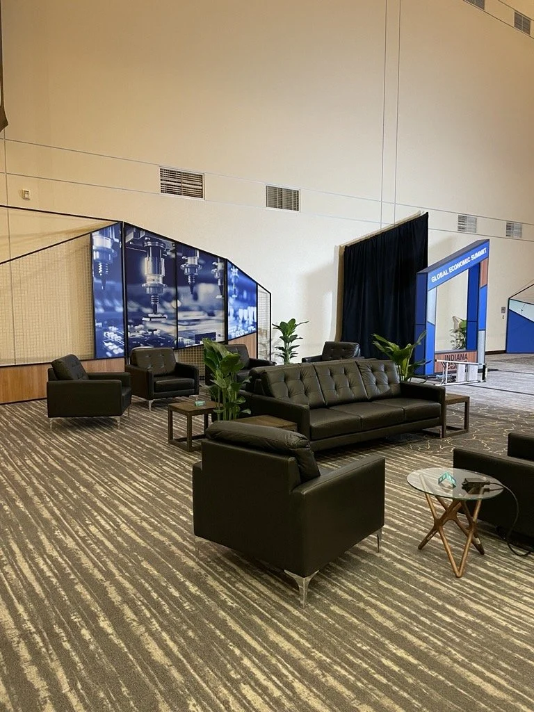 A spacious conference or waiting area with black leather sofas and armchairs, small side tables with plants, a patterned carpet, and large digital screens displaying machinery images, with a sign reading "Global Economic Summit" in the background.