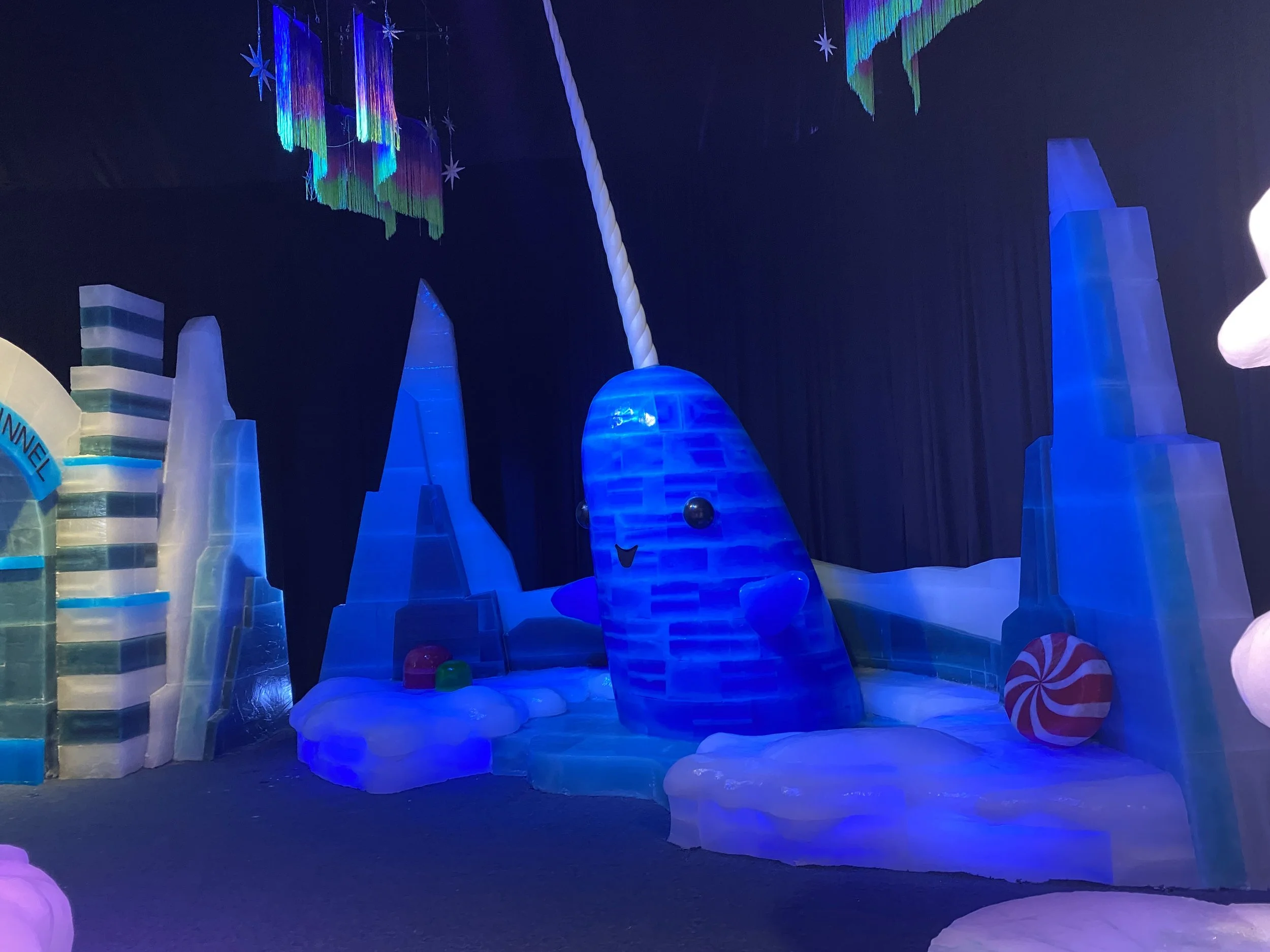 Colorful ice castle with glowing blue and white ice structures, decorations resembling stars and chimes hanging from the ceiling, and large candy-like props on the floor, creating a festive winter scene.