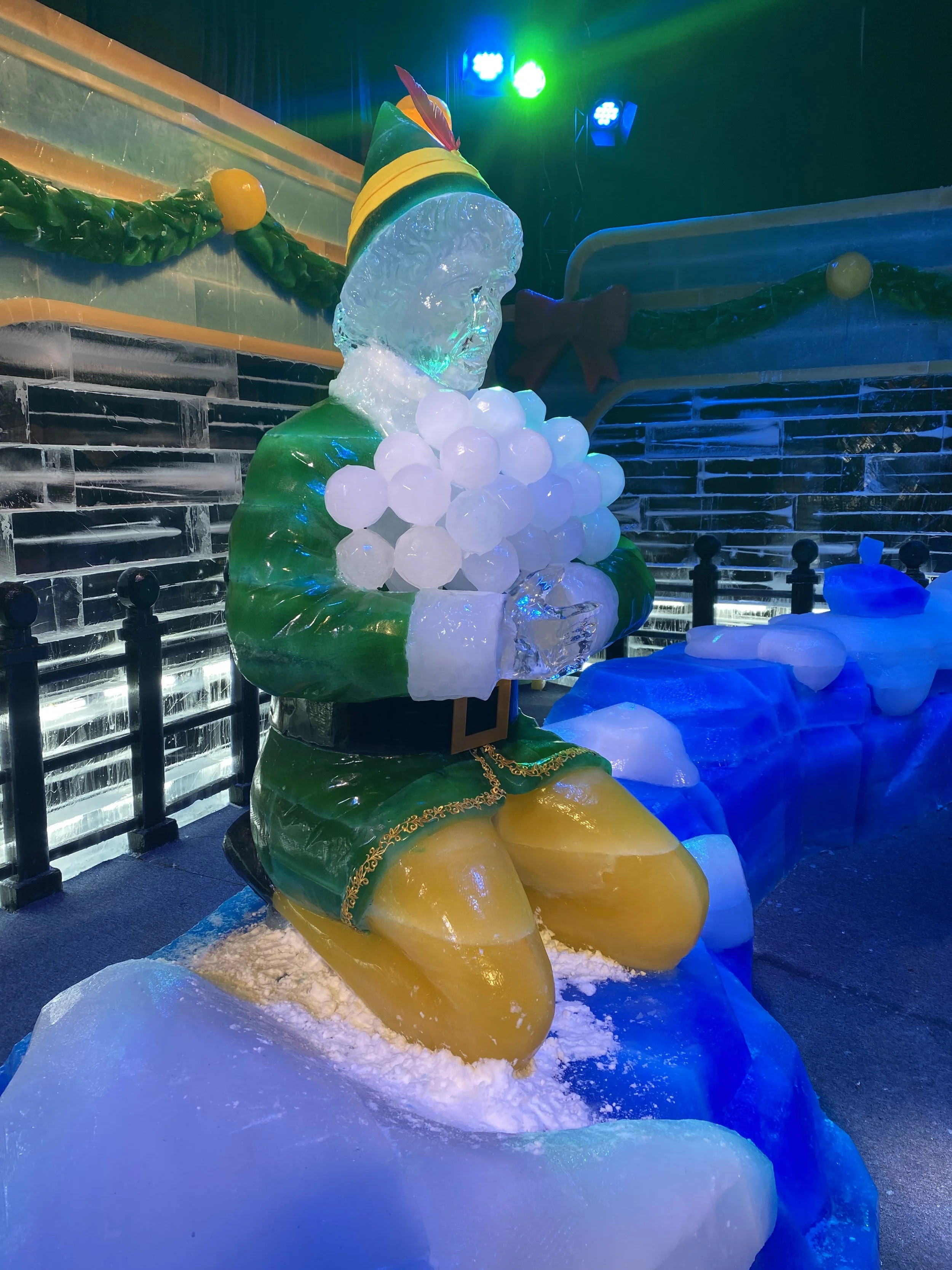 Ice sculpture of a seated elf with a colorful hat and a large bunch of ice balls in his lap, illuminated with multicolored lights.