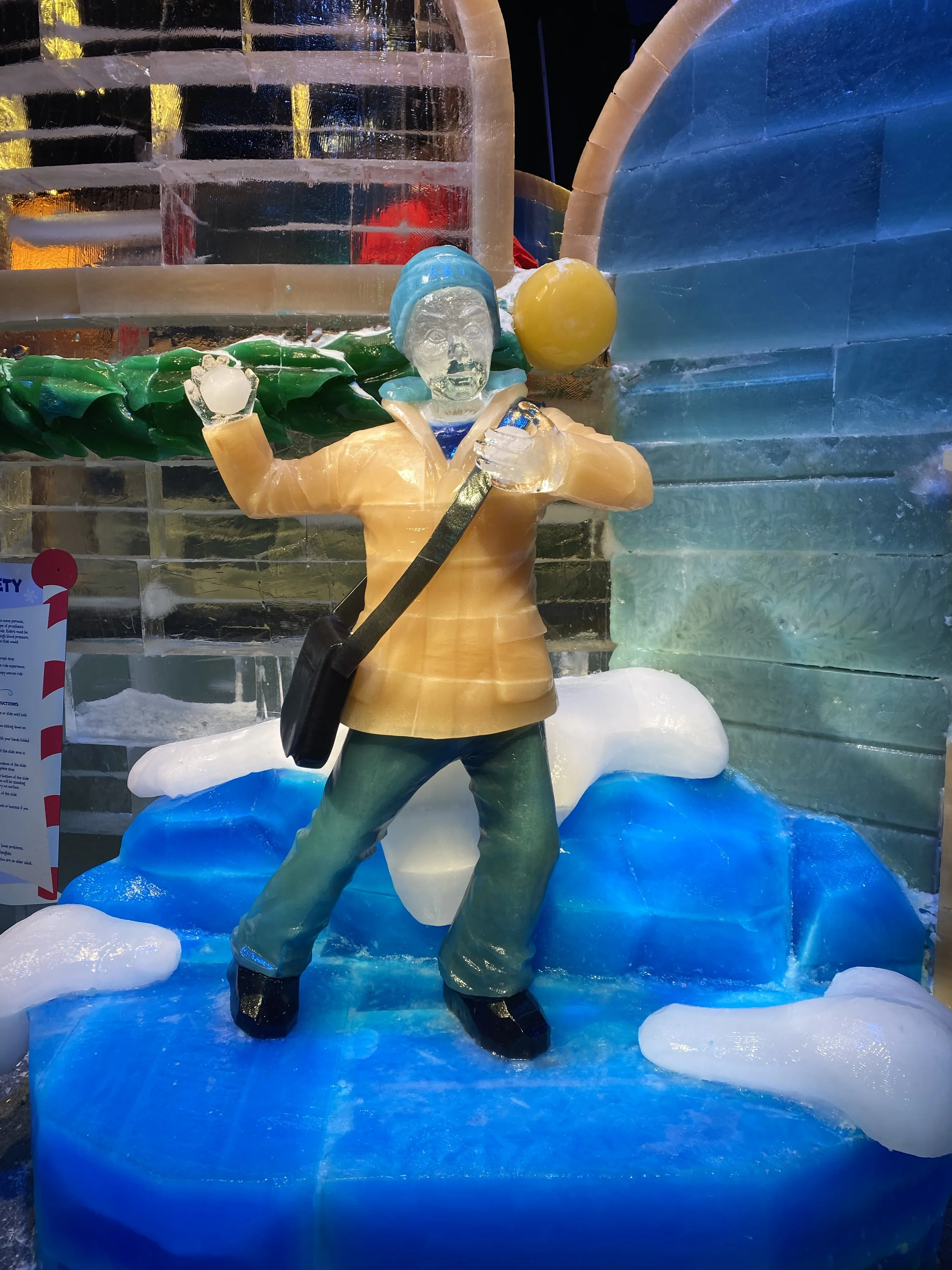 Ice sculpture of a person in winter clothing holding a fishing pole and a yellow ball, standing on an icy surface with ice blocks in the background.