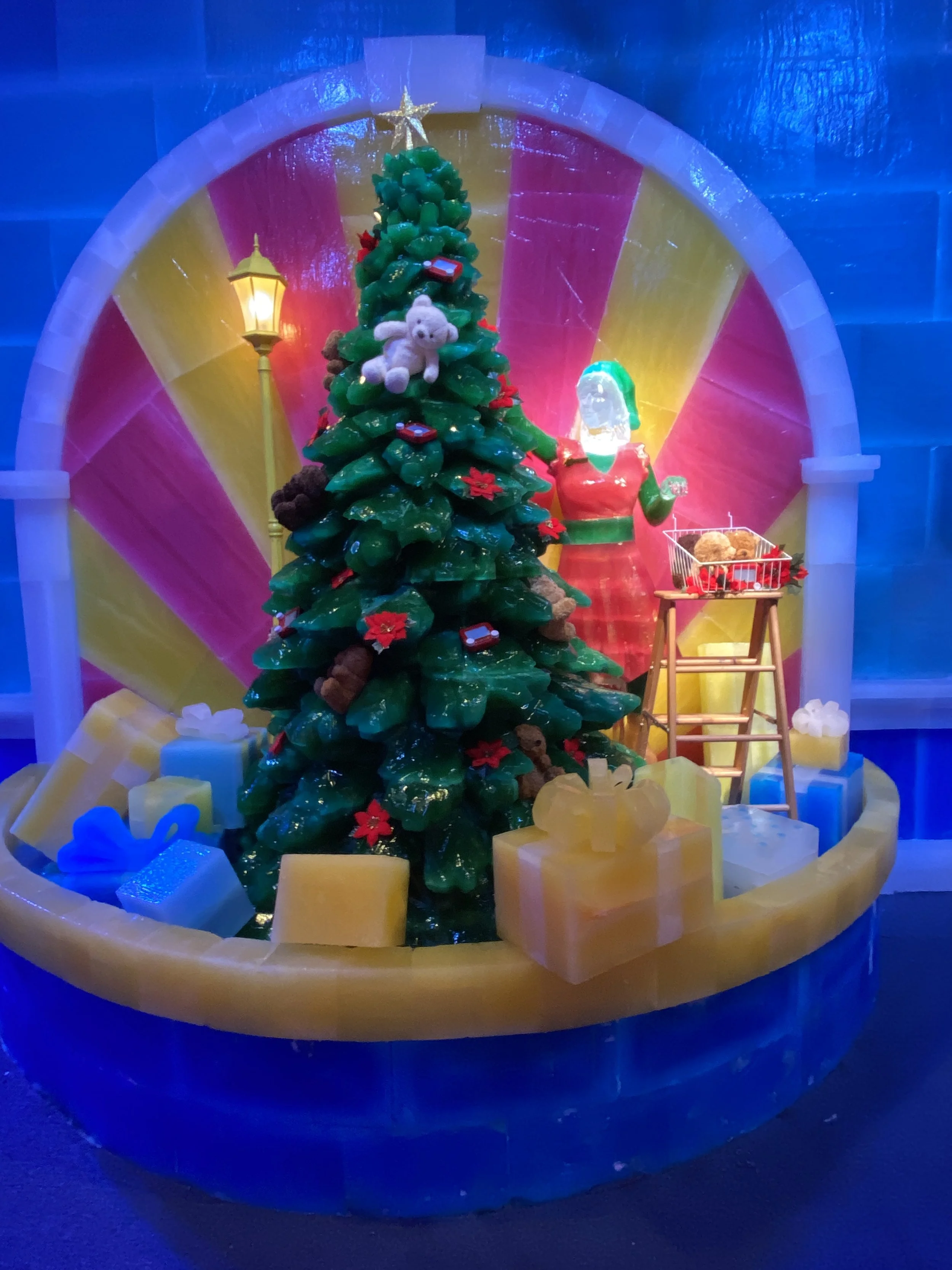 Decorative Christmas scene featuring a Christmas tree decorated with plush toys and small gifts. There is a lit street lamp, a small table with a gift basket, and large wrapped presents at the base of the scene. The background includes colorful, illu