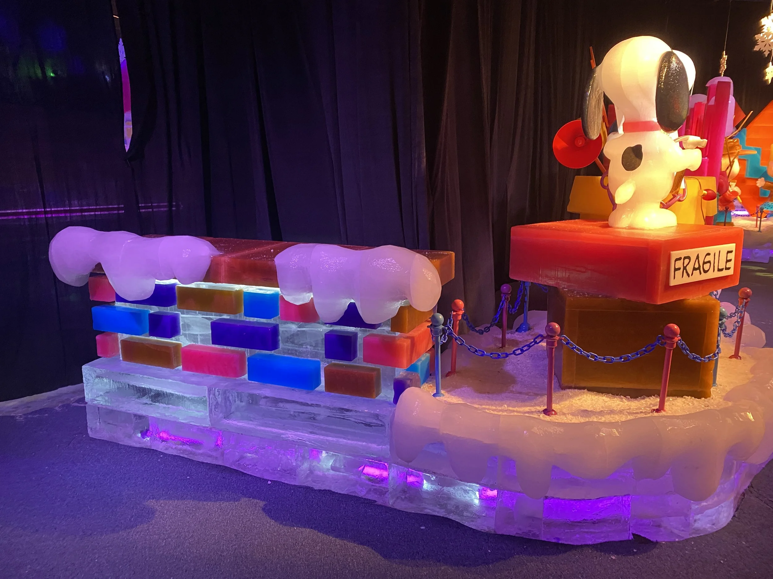 Colorful ice sculpture display with characters and blocks and a sign that says "Fragile," illuminated with purple and yellow lighting.