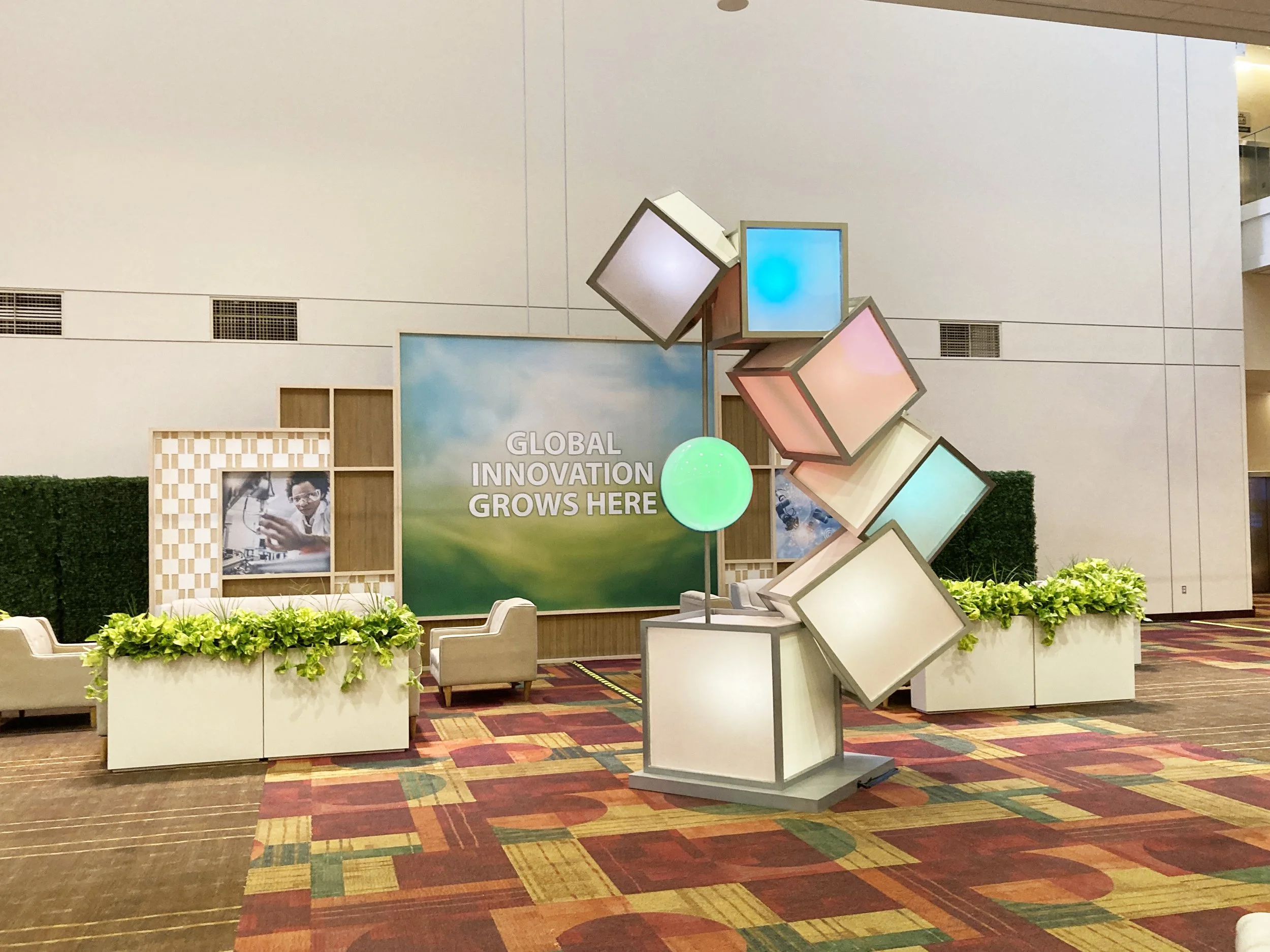Display at a trade show or conference with colorful cubes and a sign that says "Global Innovation Grows Here". Includes seating and greenery.