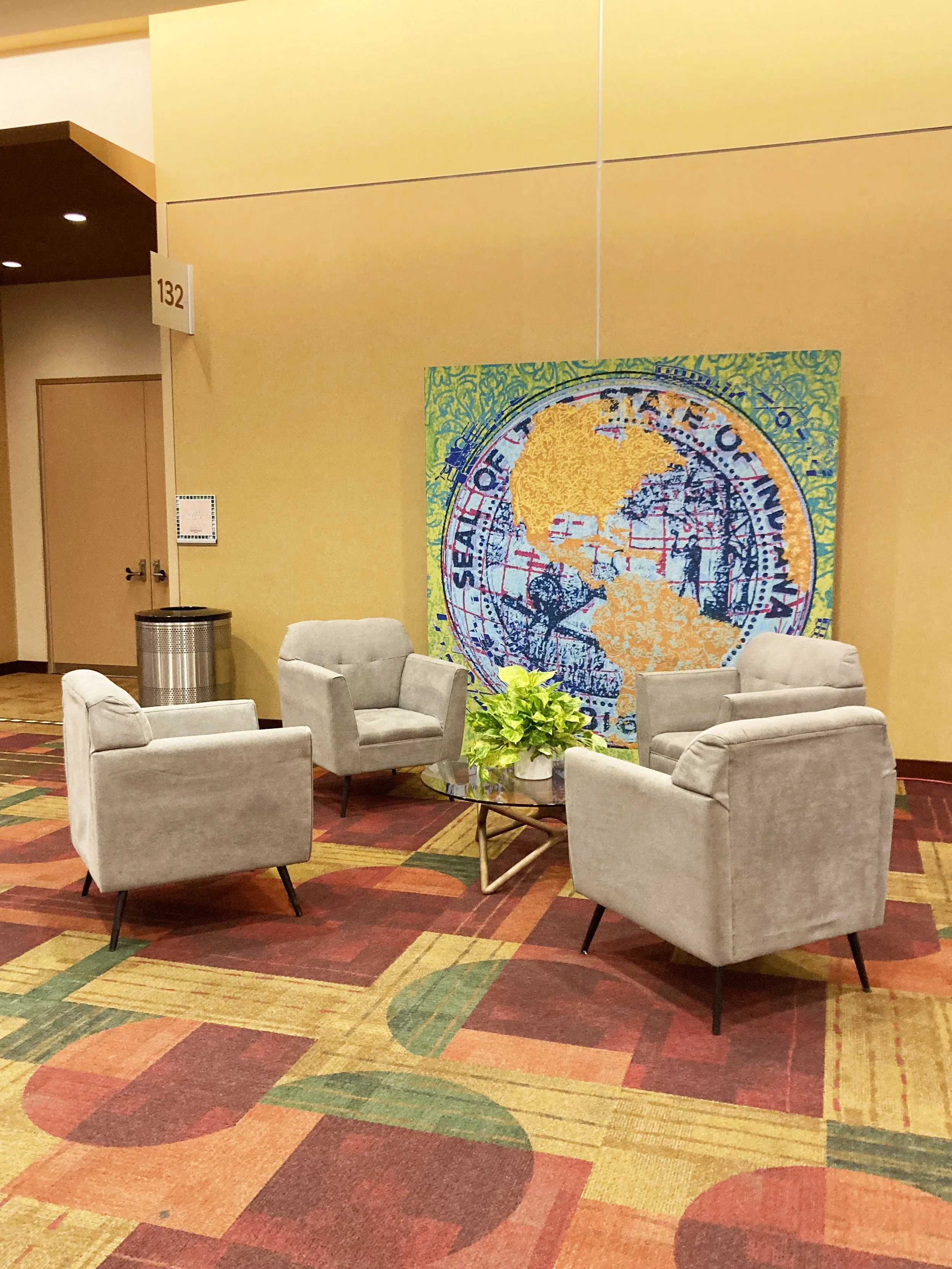 A colorful hotel lobby seating area with four light gray armchairs arranged around a small glass-top table with a green potted plant. In the background, a large North American map poster is partially visible on the wall. The floor has a multicolored 