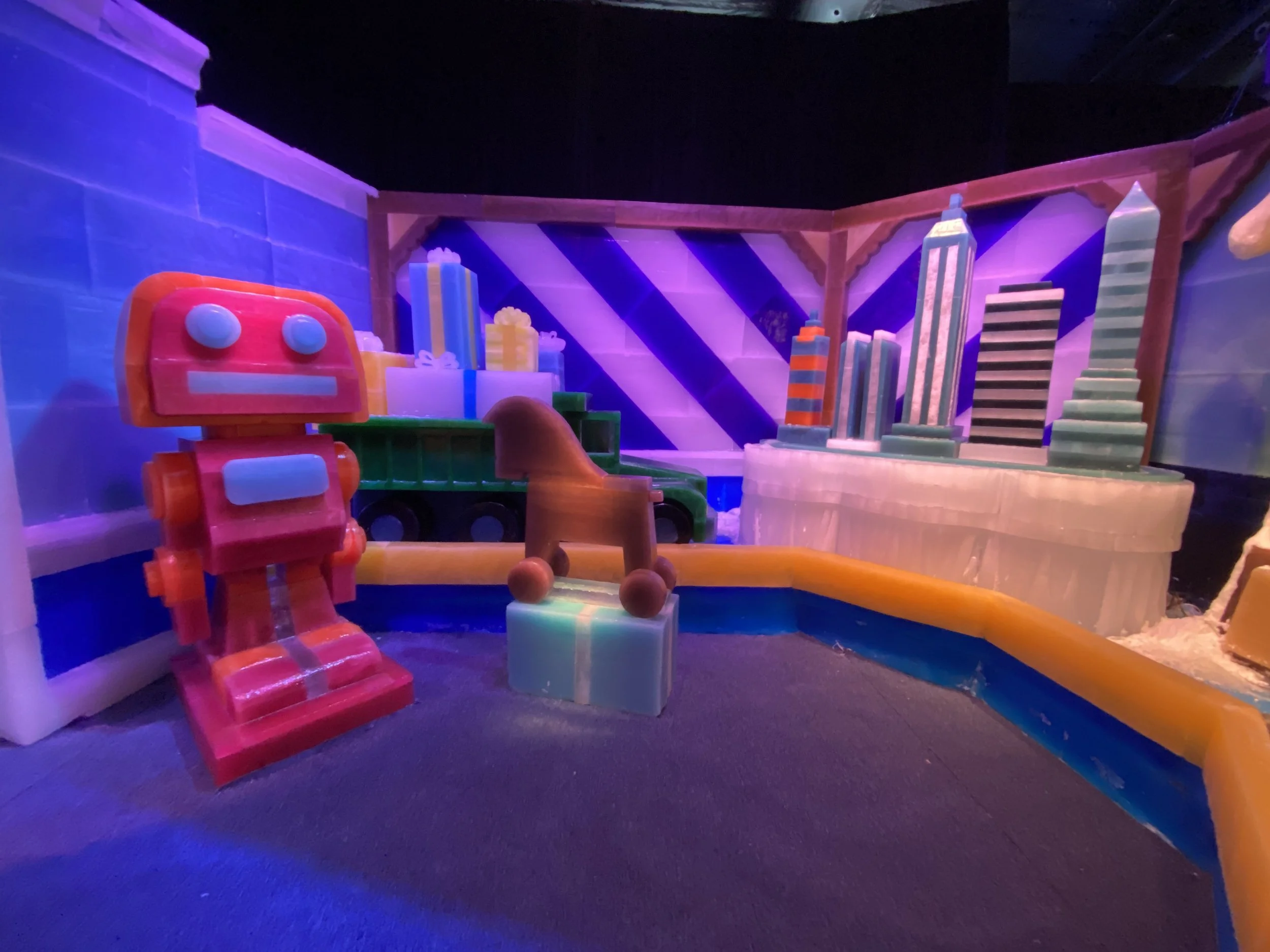 Colorful indoor playground with a red slide, soft pink and orange play structures, and large cityscape murals on the walls, illuminated with vibrant purple and blue lighting.
