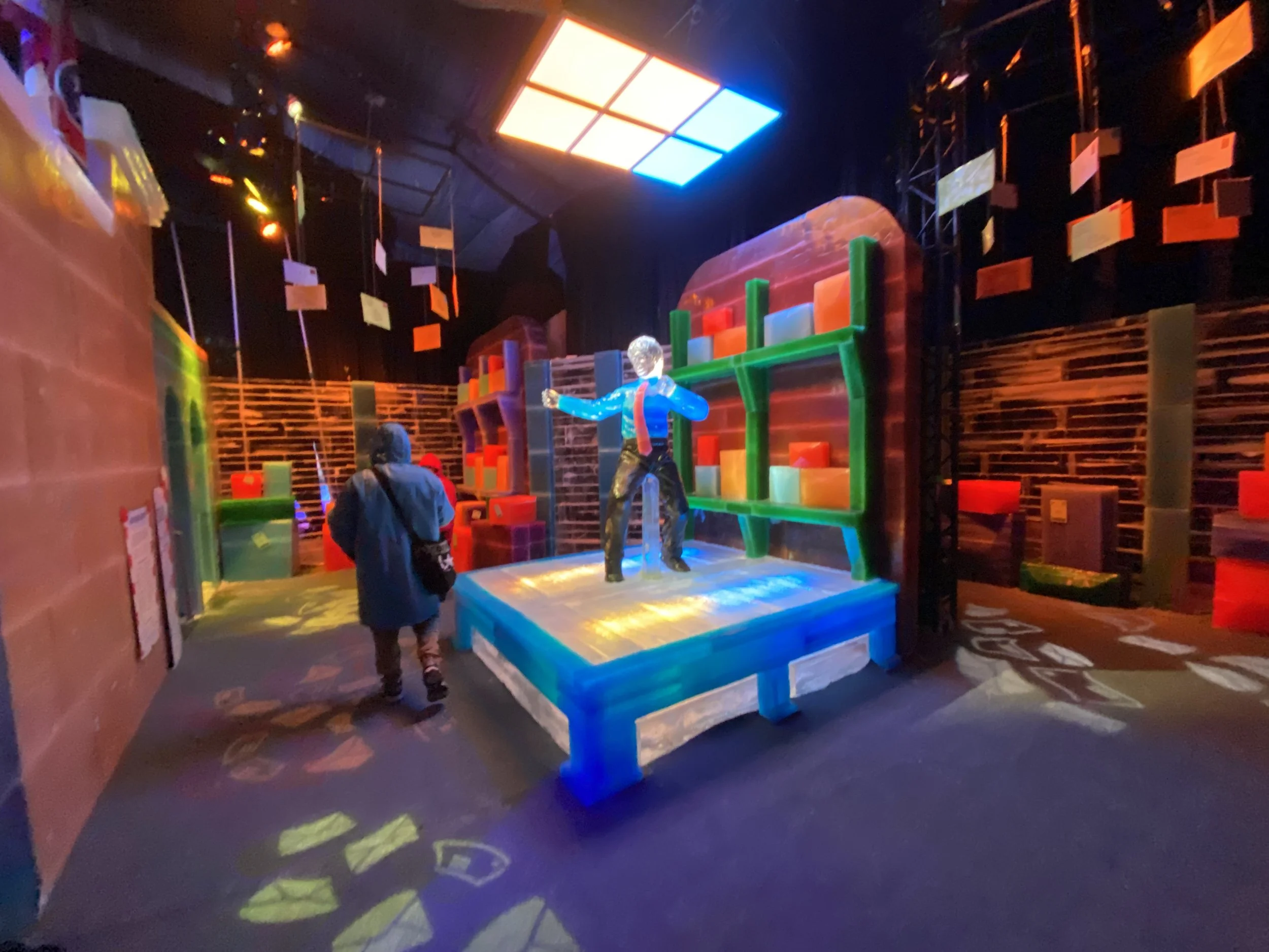 Colorful, illuminated indoor exhibit area with geometric and abstract structures, including a mannequin on a platform, and walls decorated with bright, neon-like lights and hanging signs.