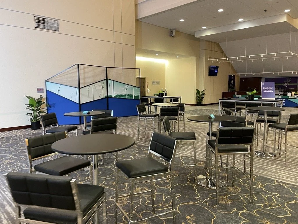 Modern hotel lobby with black chairs and round tables, plants, and a children's play area with a padded enclosure and blue panels.