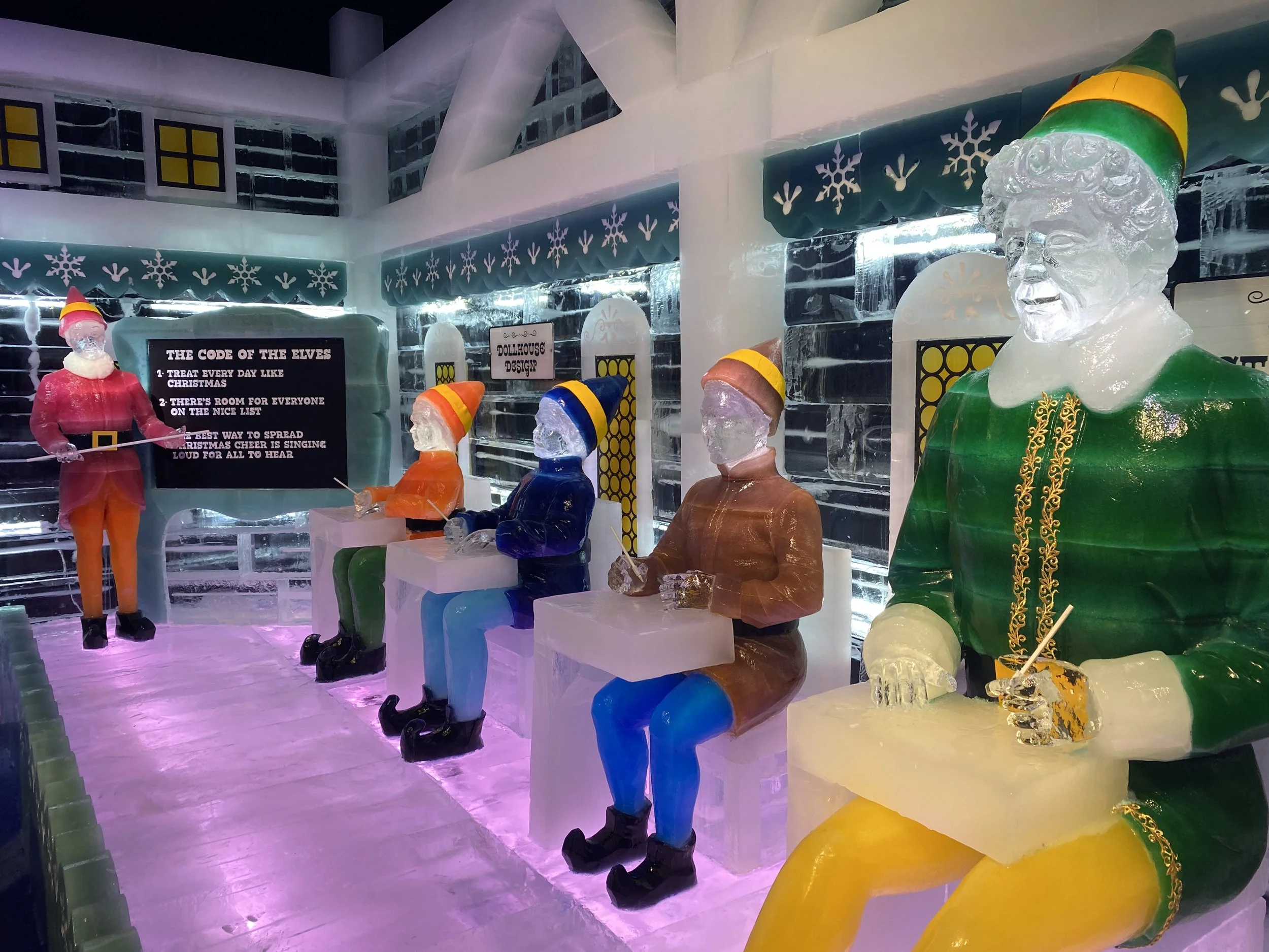 Ice sculptures of elves dressed in colorful clothing, sitting and standing, with a large ice Santa Claus figure in the background, decorated for Christmas.