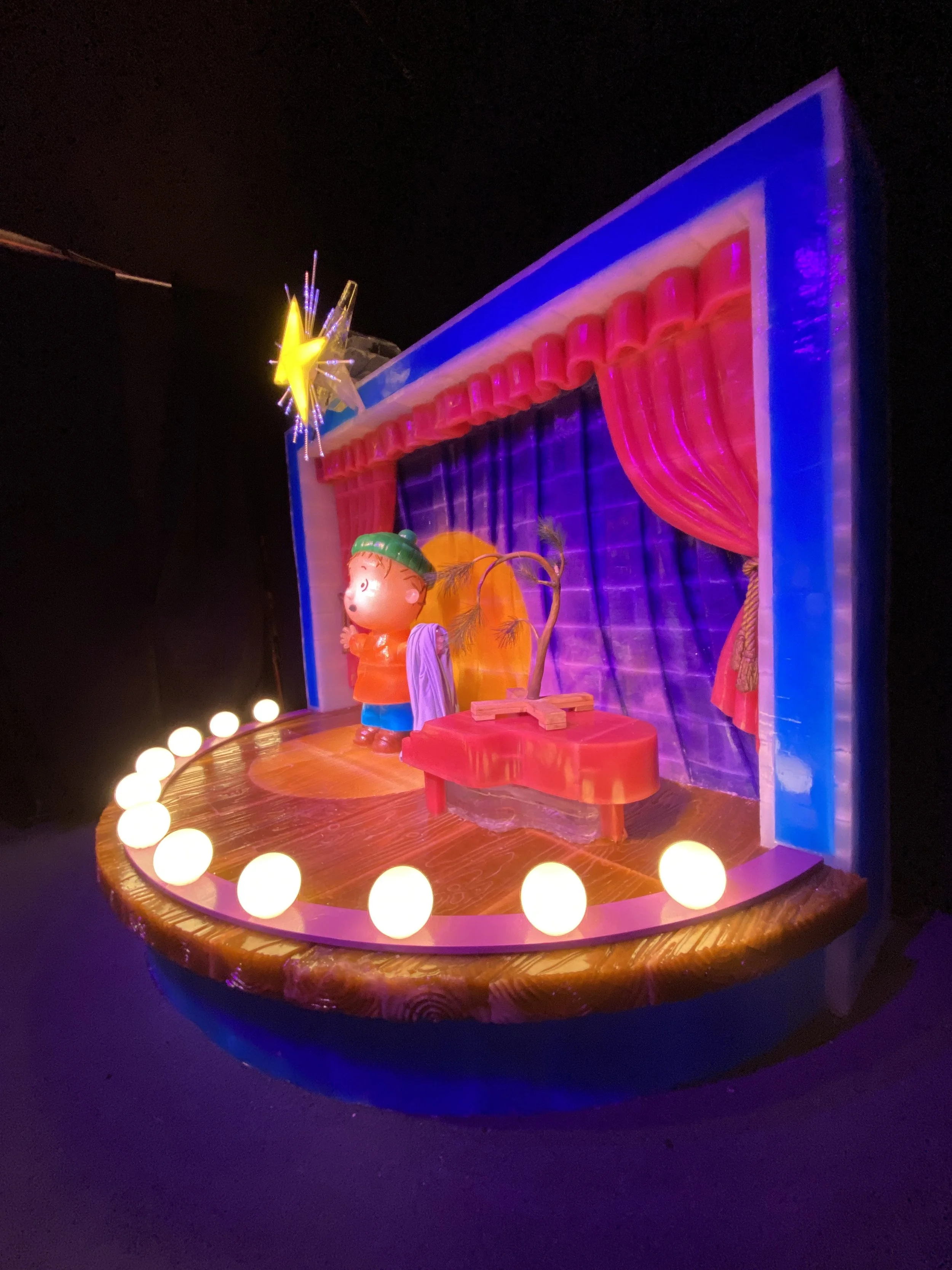 Colorful carnival-style puppet stage with pink curtains, a doll with a green hat, a small tree, orange pumpkins, surrounded by round lights in a semi-circle.