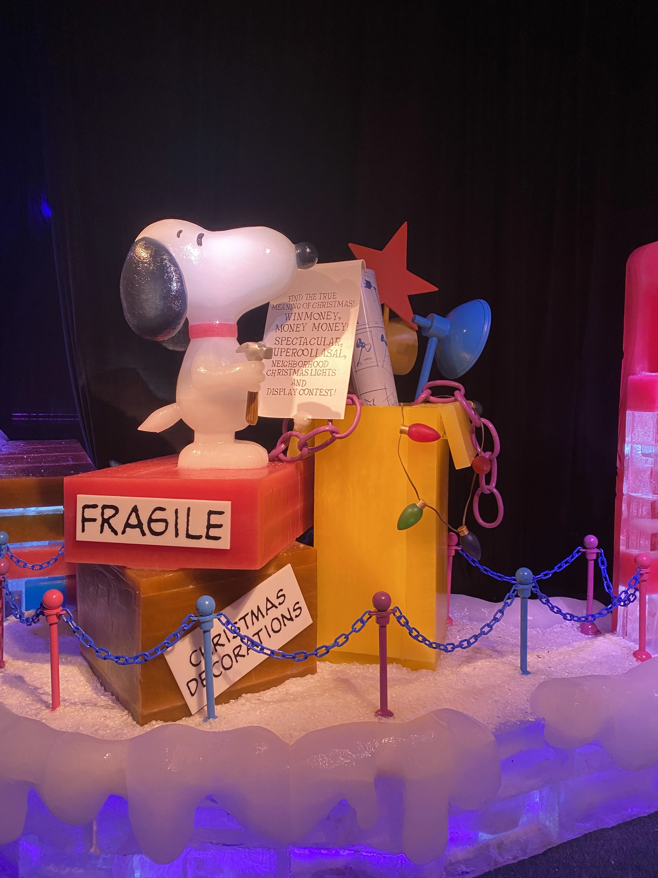 Decorative holiday display with Snoopy holding a Christmas letter, surrounded by colorful props, lights, and signs reading 'FRAGILE' and 'CHRISTMAS DECORATIONS' in a winter-themed setting.