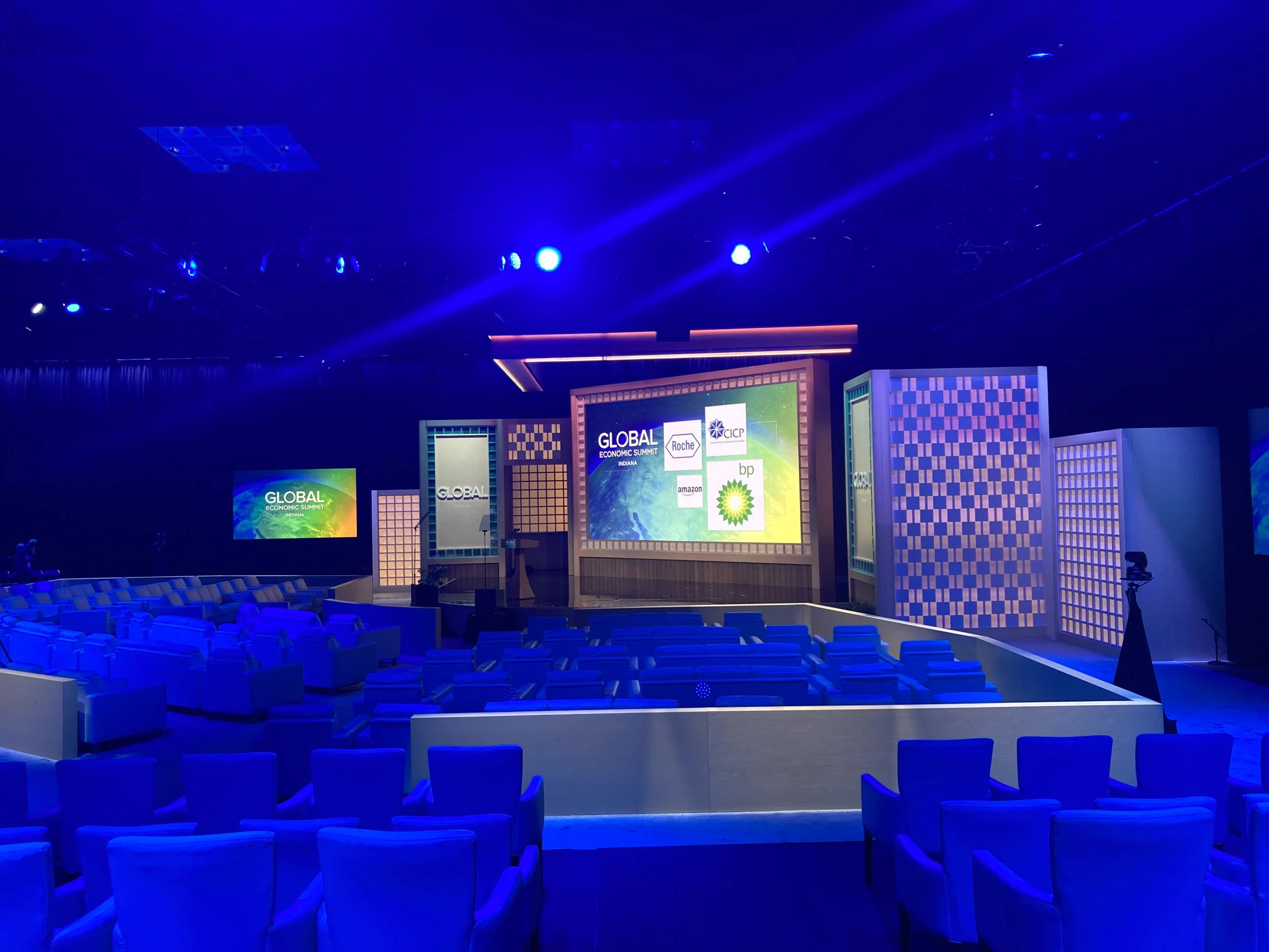 Empty conference hall with blue lighting, rows of chairs, and a stage with multiple screens for the Global Economic Summit Indiana.