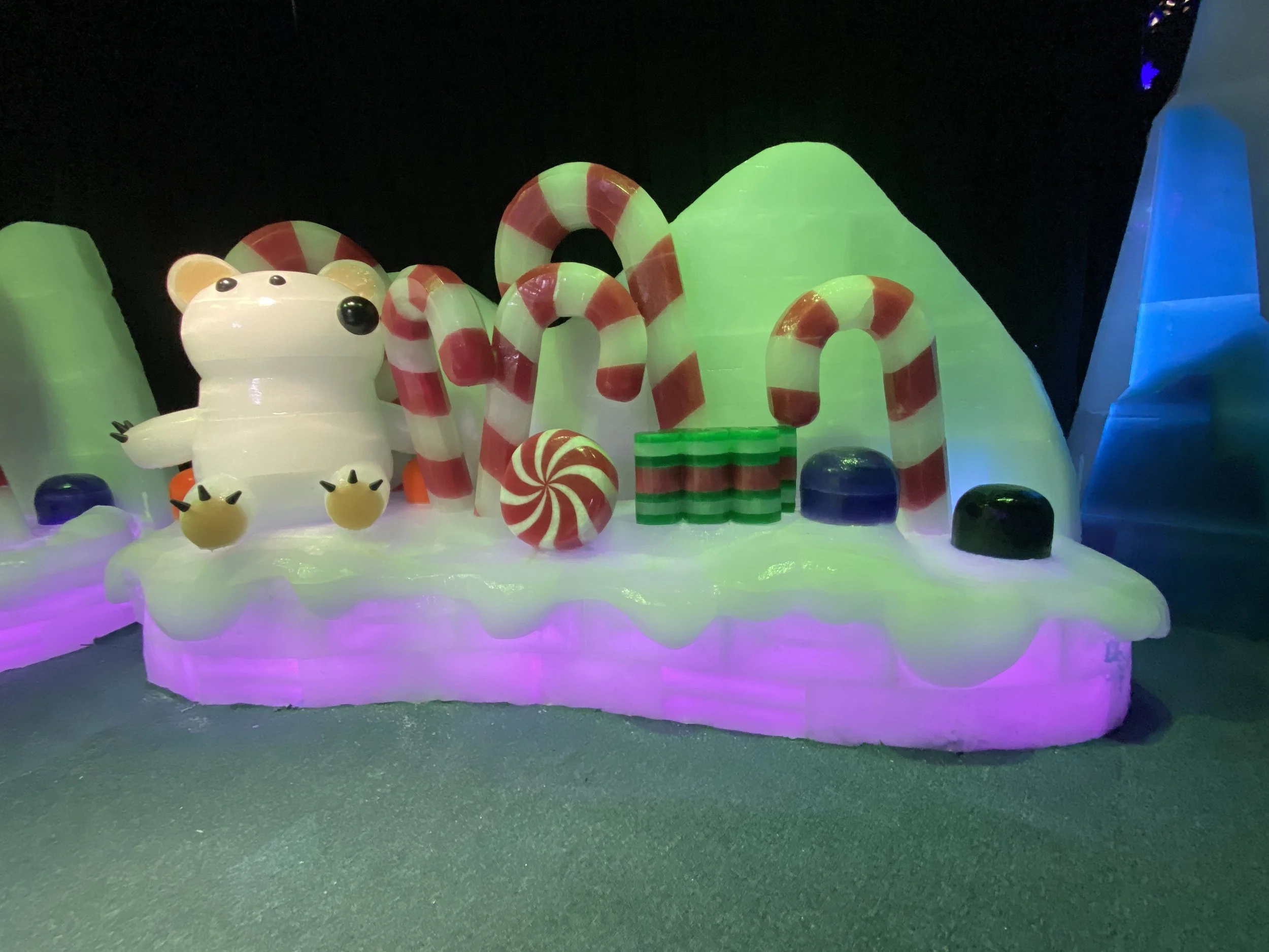 Colorful holiday-themed display with a glowing polar bear, striped candy canes, and various brightly colored objects on a glowing base, illuminated in a dark setting.