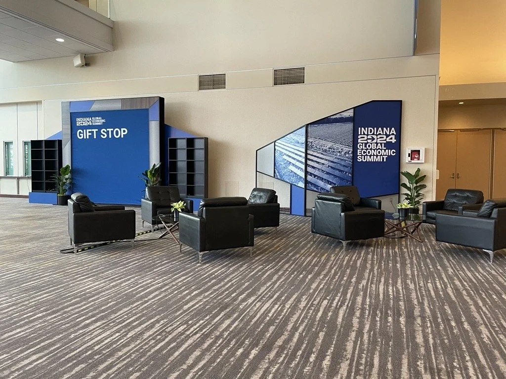 Indoor seating area with five black leather chairs, small tables, and potted plants, near a large digital display with the text "Gift Stop" and "Indiana 2024 Global Economic Summit."