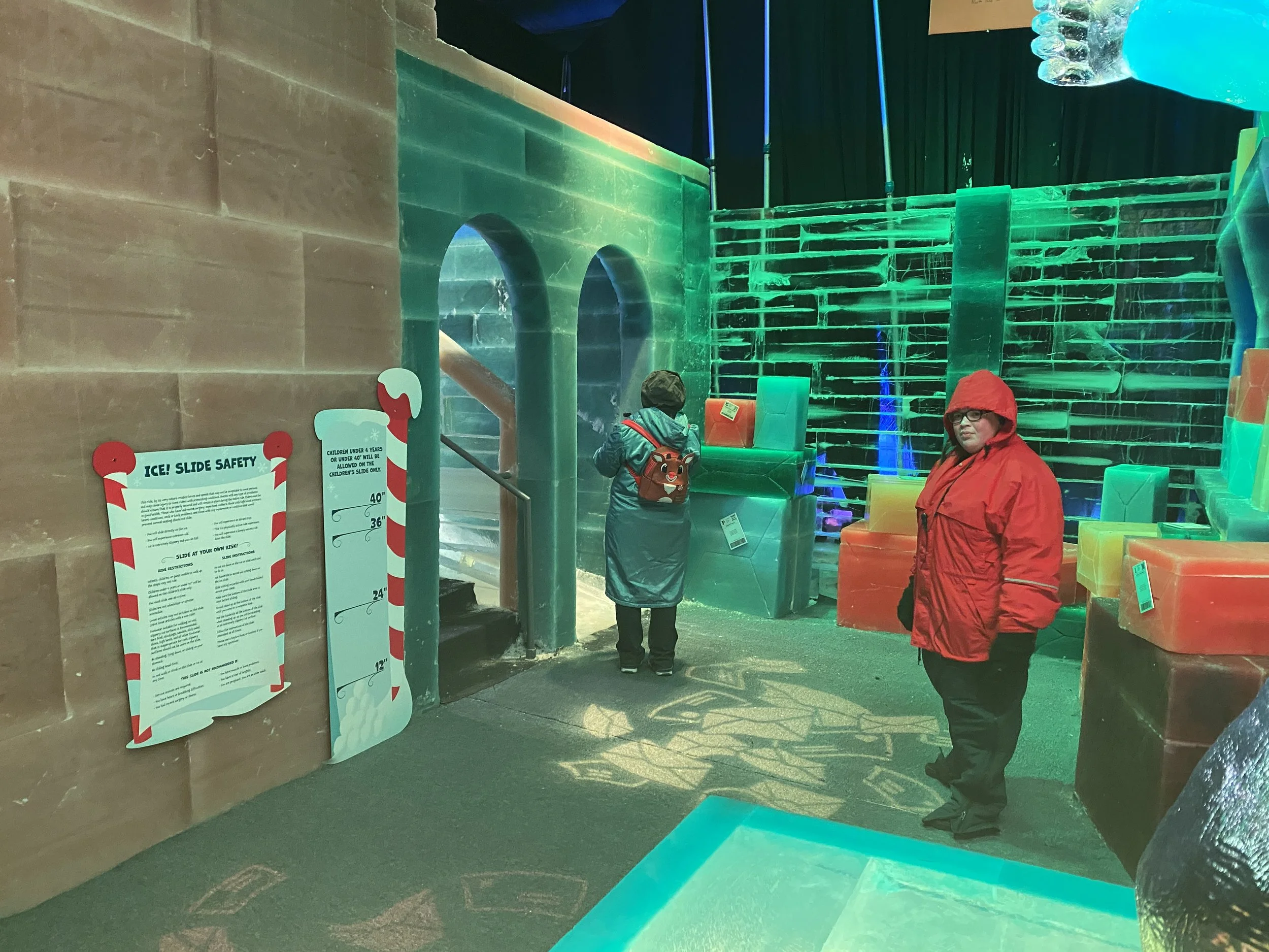 Inside an ice-themed exhibit, two children and an adult are present. The decor includes large, colorful ice sculptures and structures, illuminated with green and blue lighting. A sign displays ice slide safety instructions, indicating slide heights a