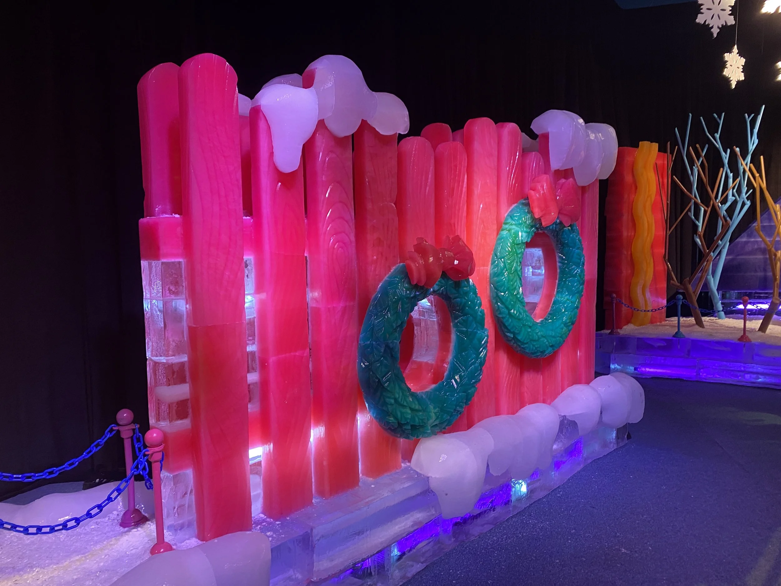 Colorful ice sculpture display with pink, white, green, and yellow elements, surrounded by snow and blue chain barriers, illuminated by purple lights.