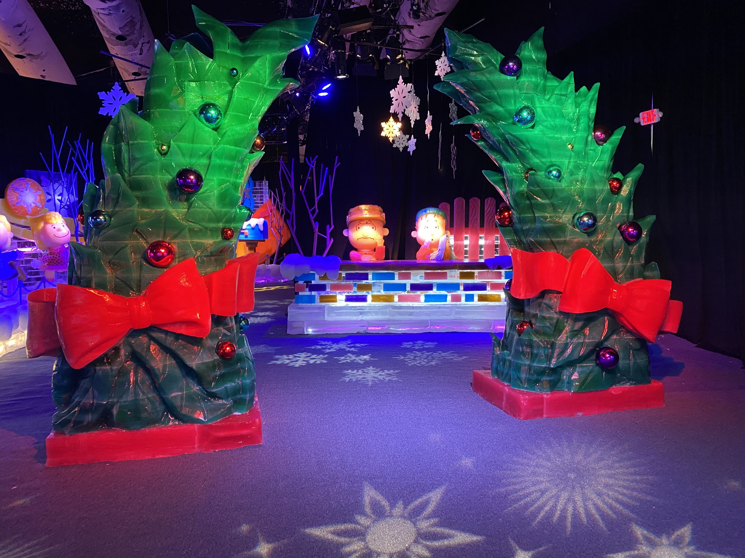 Colorful holiday display featuring large green artificial holly leaves with multicolored ornaments, snowflake decorations hanging from the ceiling, and various cartoon character figures in a dark room, illuminated by vibrant lights.
