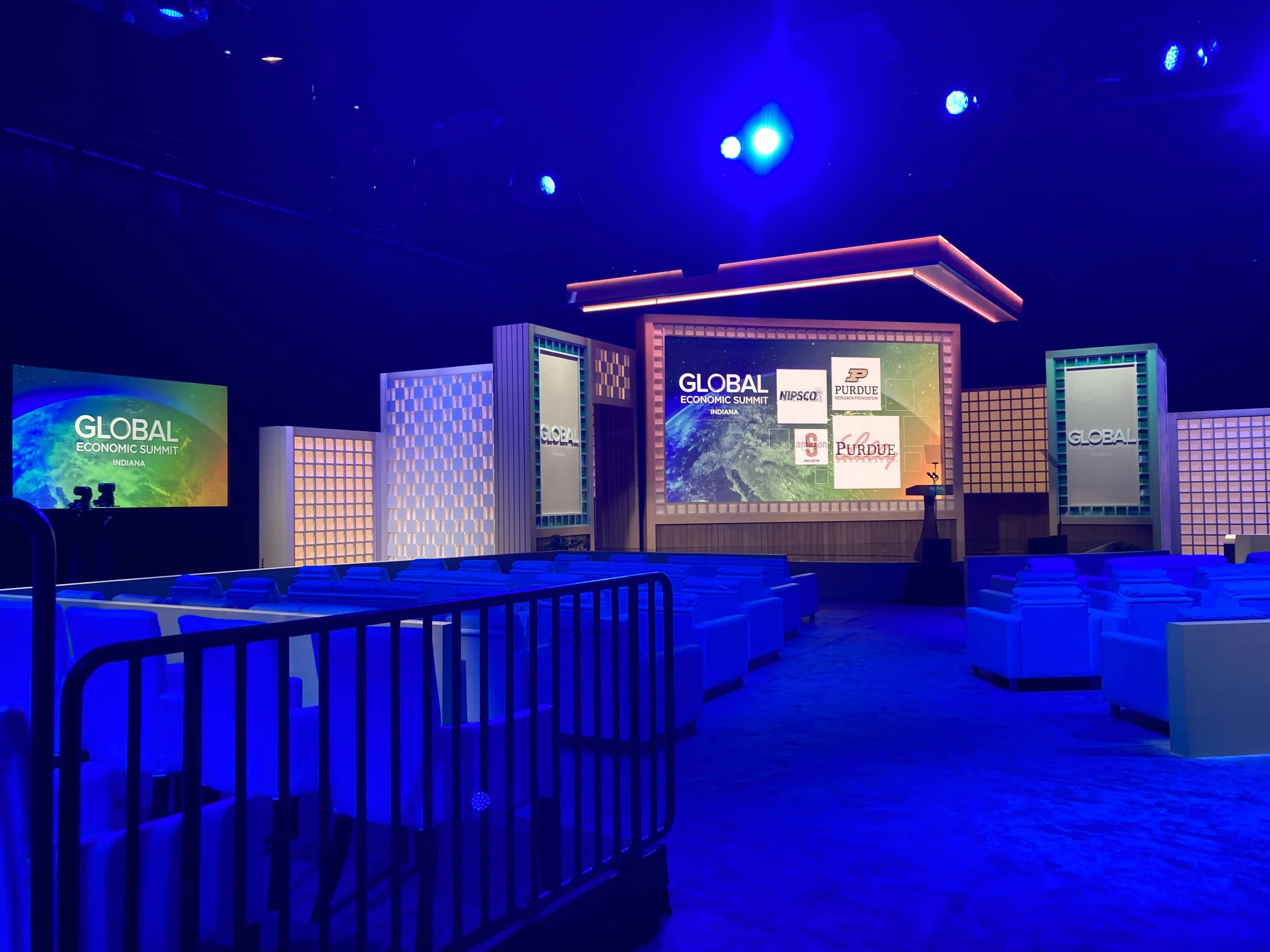 Stage setup for the Global Economic Summit with large screens displaying the event's name and logos, illuminated by blue and purple lighting, with empty chairs in the foreground.
