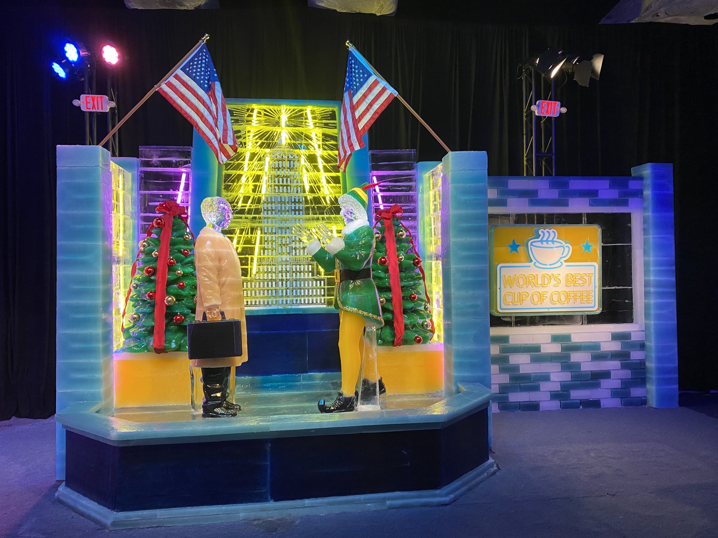 Ice sculpture display featuring two figures, Christmas trees with red ribbons, American flags, and a yellow neon light backdrop. Sign reads "World's Best Cup of Coffee".