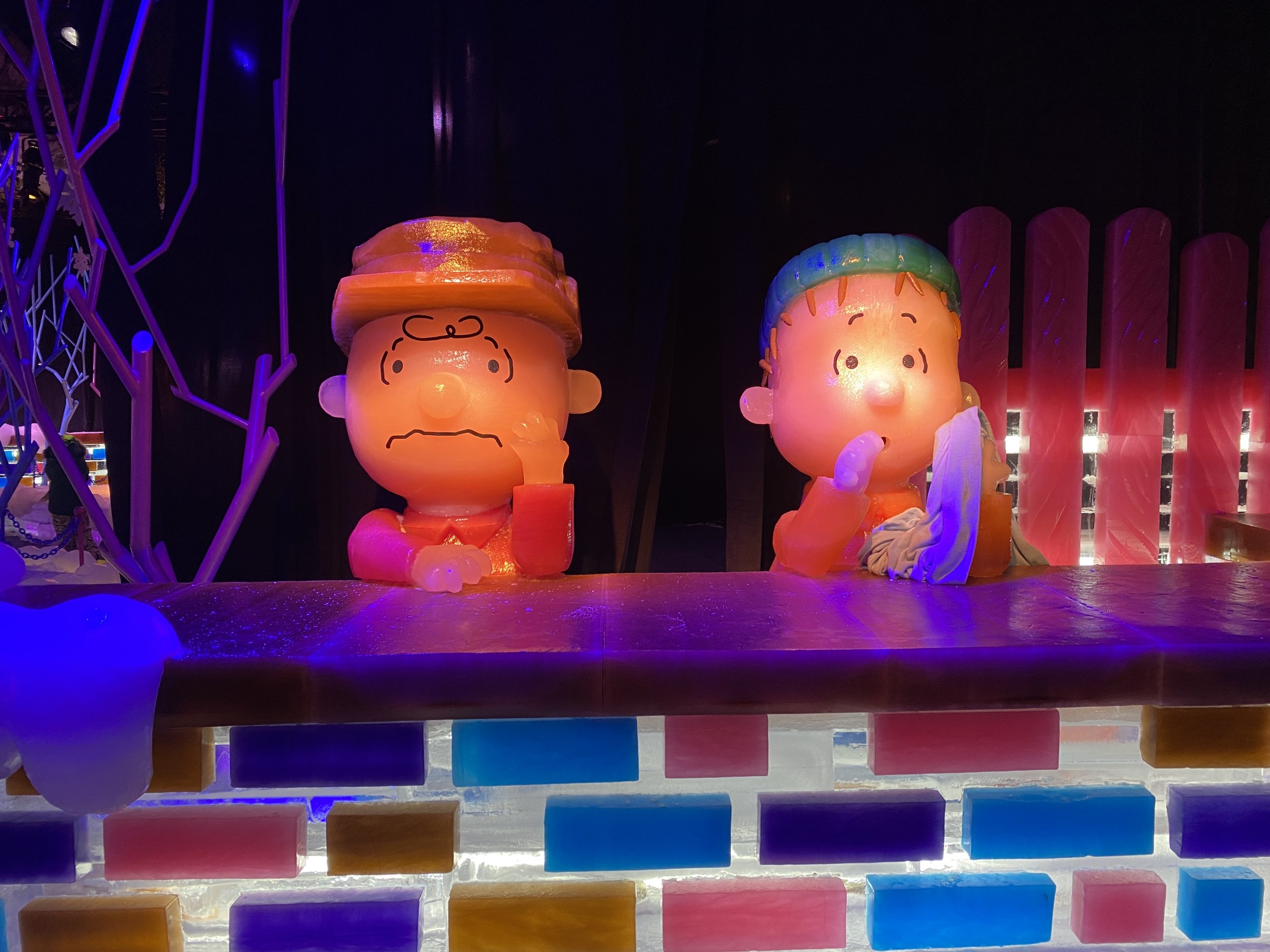 Colorful illuminated sculptures of Charlie Brown and Linus from Peanuts during a night exhibit, with rainbow-colored ice blocks and artistic decorations around.