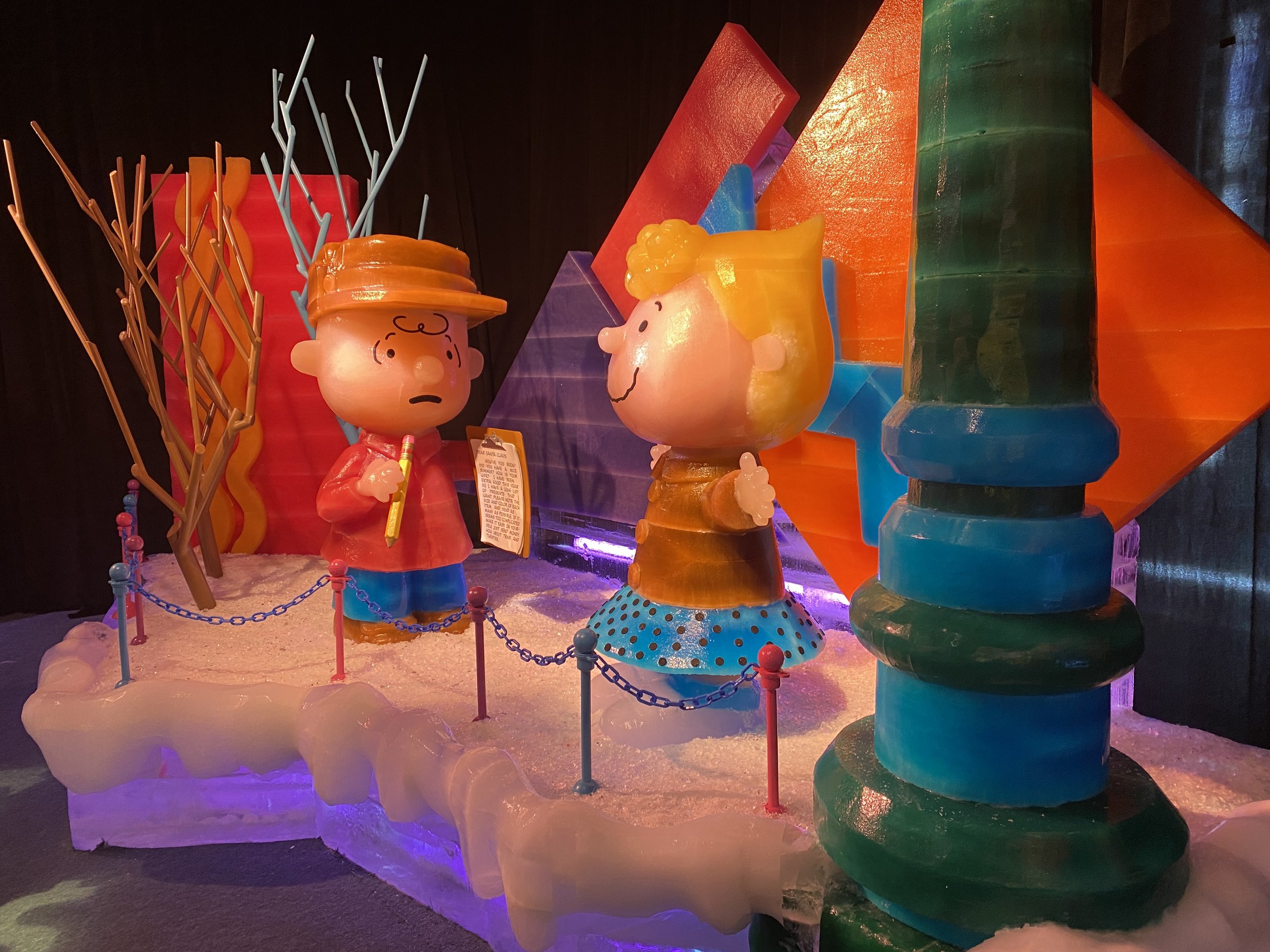 Colorful cartoon-style display with characters and shapes, including a boy with a hat and a girl with yellow hair, surrounded by bright geometric structures and a chain fence.