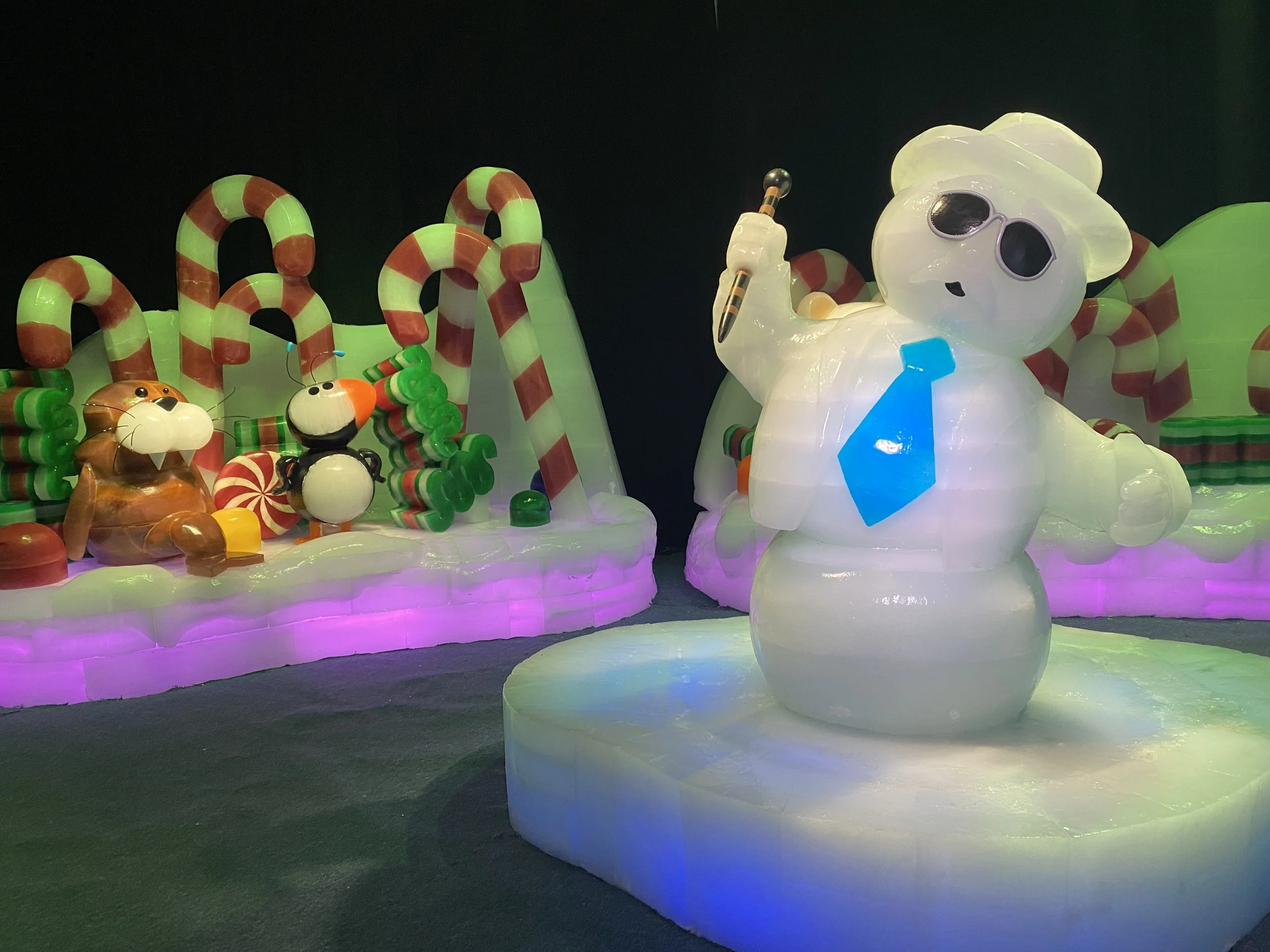 Colorfully lit ice sculpture display featuring a large bear wearing sunglasses and holding a pencil, with smaller animal figures and candy cane-like structures in the background.
