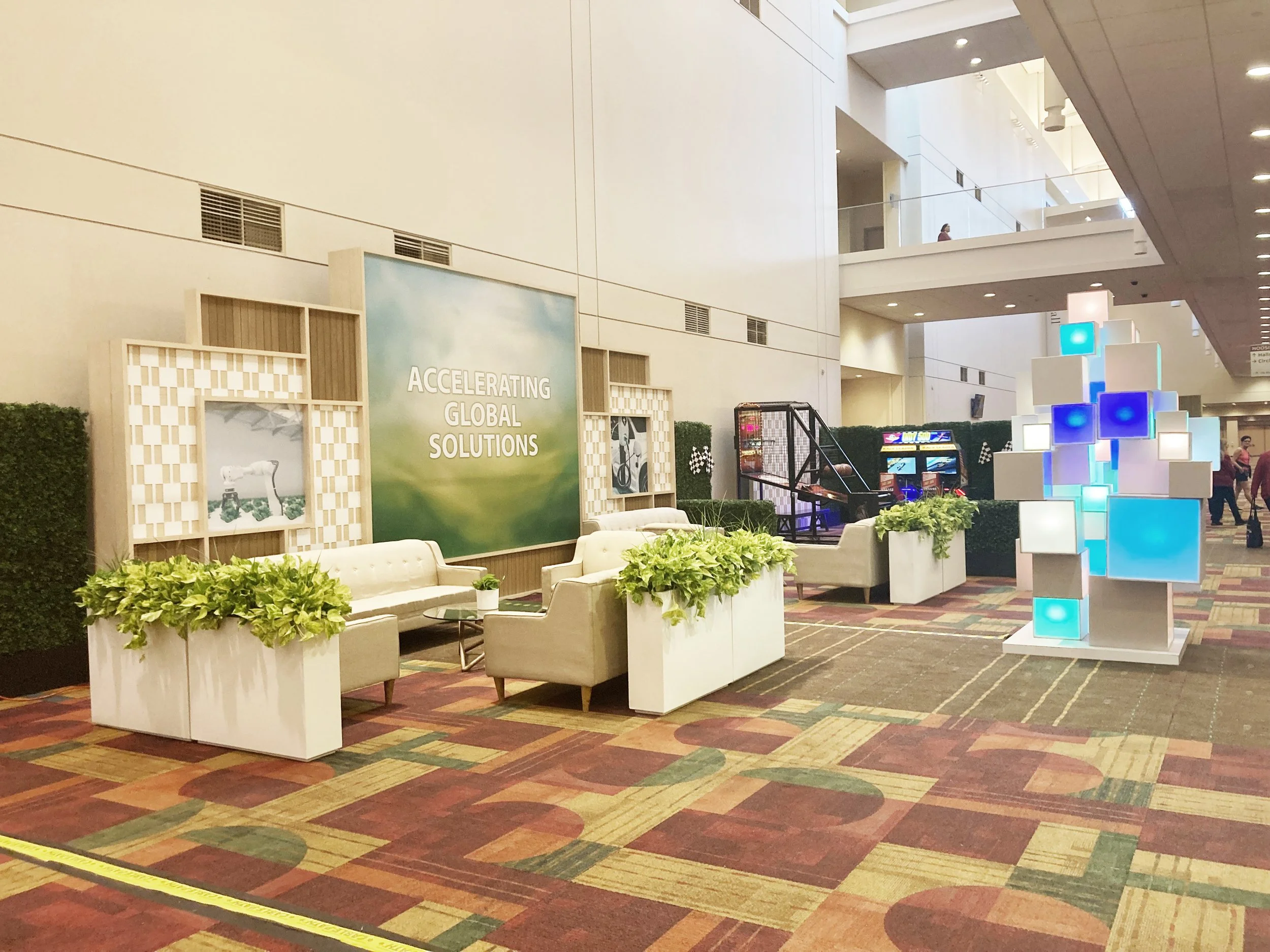 Interior of a modern building with a high ceiling, seating area with white cushioned chairs, plants, and a large sign that reads 'Accelerating Global Solutions.' There are arcade games, hanging lights, and people walking in the background.