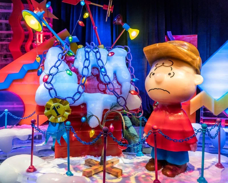 A colorful scene featuring Snoopy from Peanuts as a large sculpture with a sad expression, surrounded by chains, chains, and Christmas lights on a snowy surface, with stairs and vibrant decorations in the background.