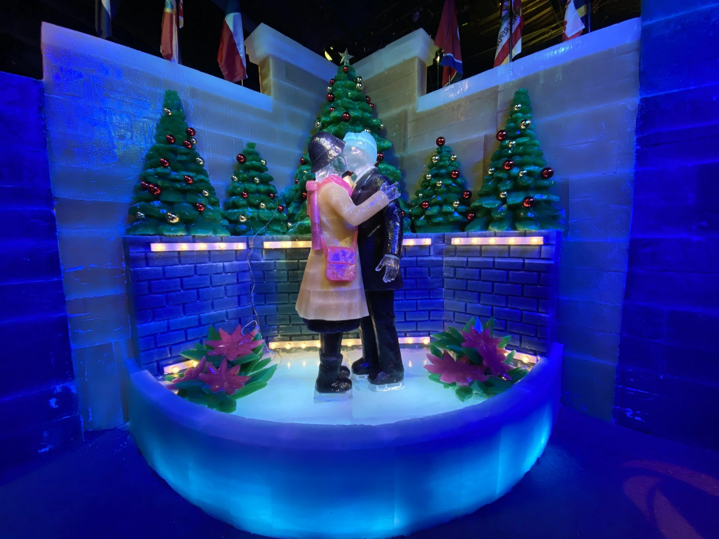 Ice sculpture of a couple kissing, surrounded by decorated Christmas trees, illuminated with blue lighting.