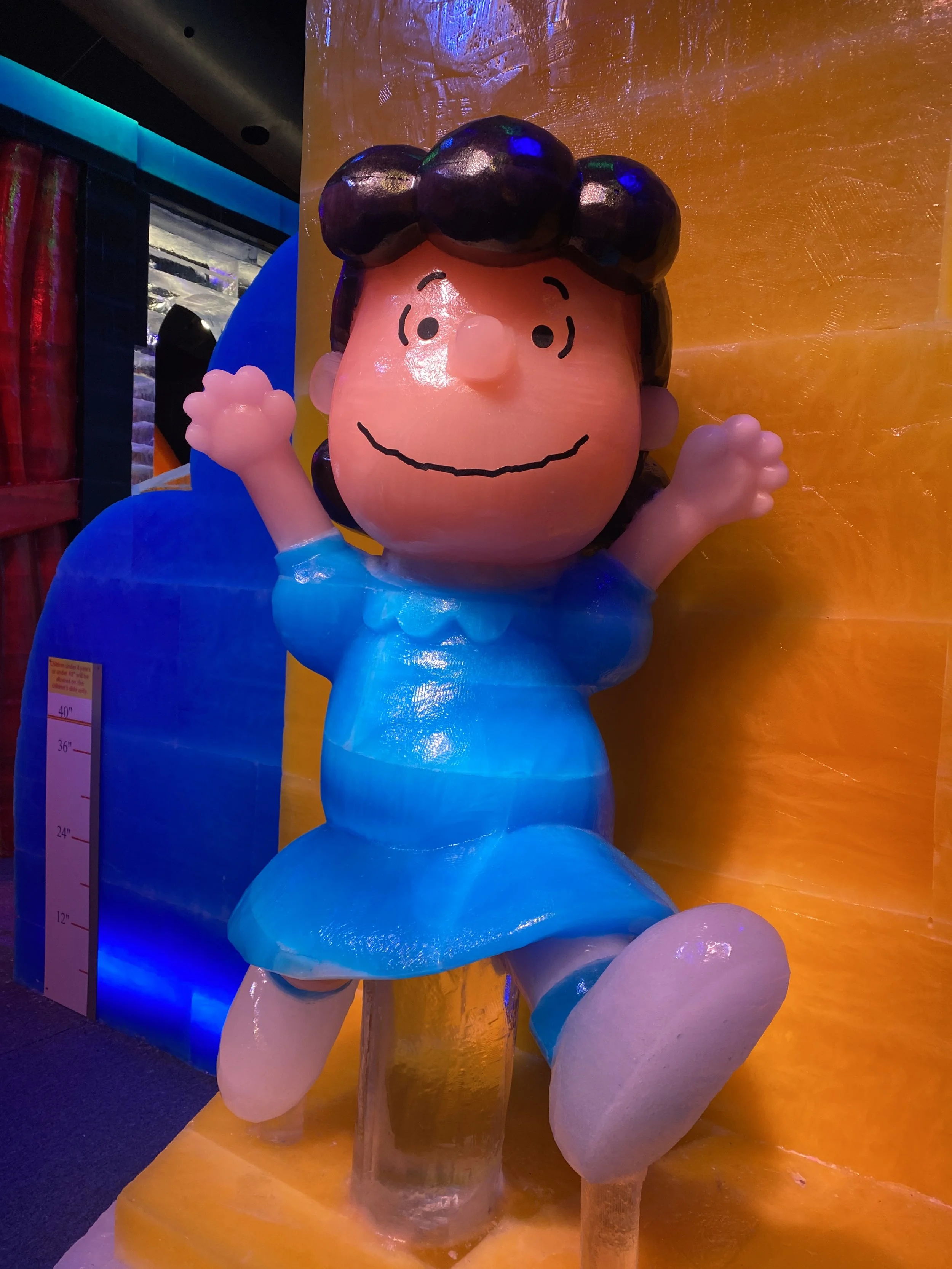 Figurine of Charlie Brown from Peanuts, with a smiling face, waving, with black hair on the head, dressed in blue, against an orange background.