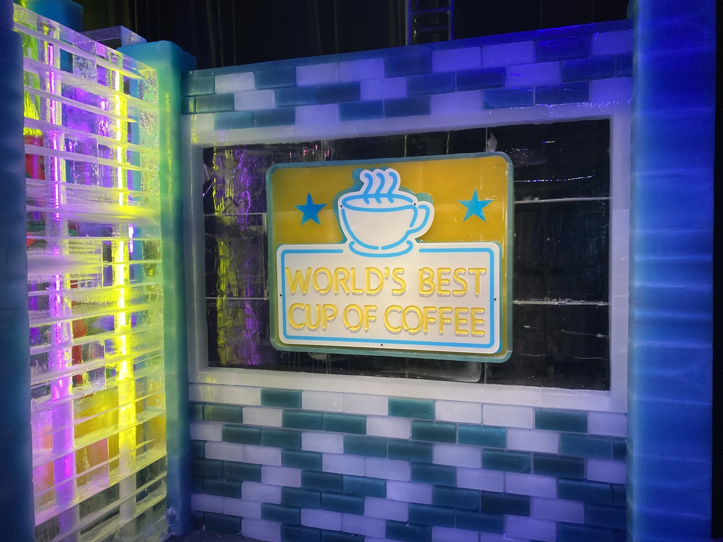 Ice sculpture with colorful LED lighting, featuring a sign that says 'World's Best Cup of Coffee' with a steaming coffee cup graphic and blue stars, set against a background of ice bricks.