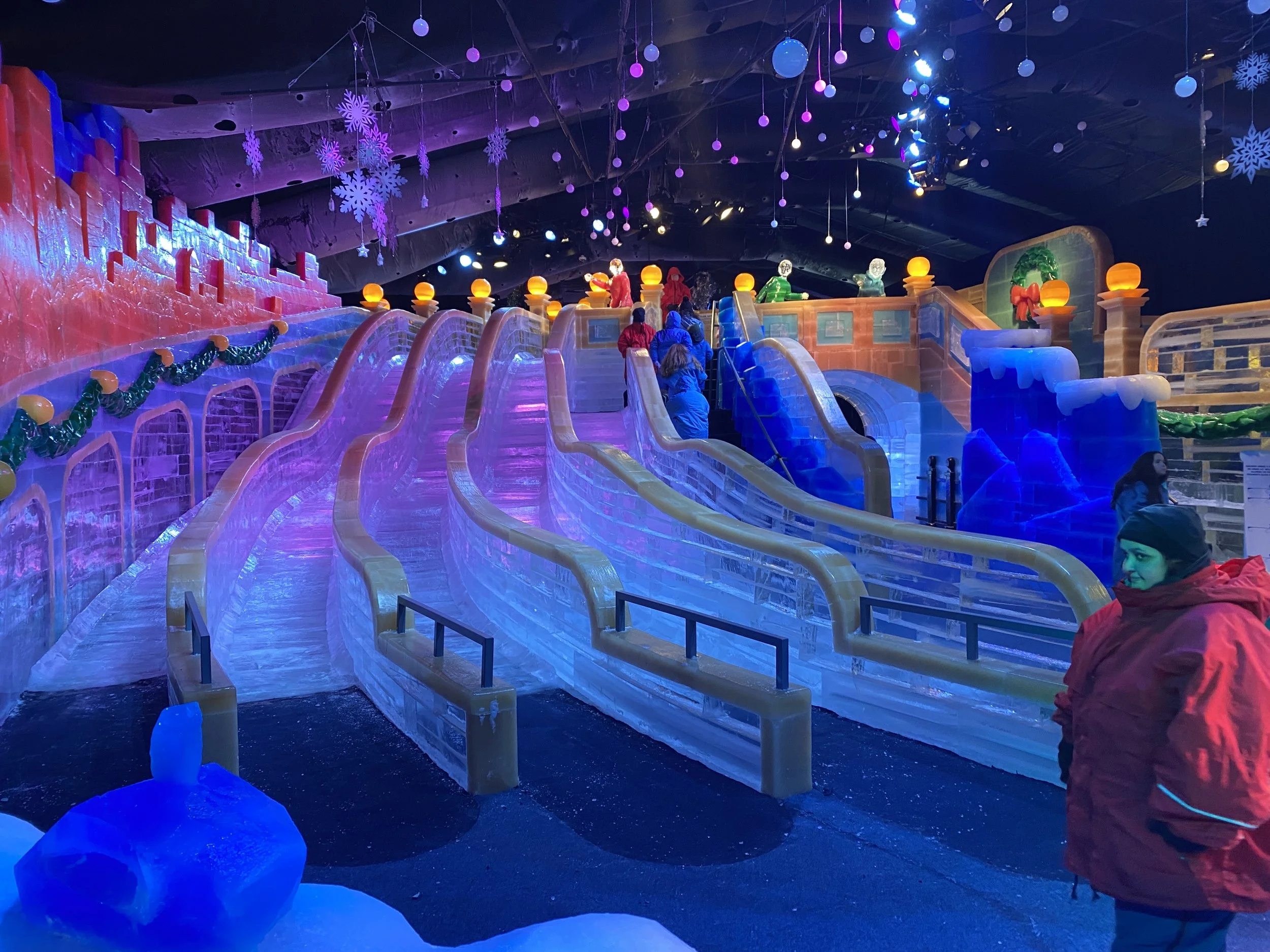 An indoor ice skating rink decorated for the holiday season with colorful lights, ice skating slides, and people skating.